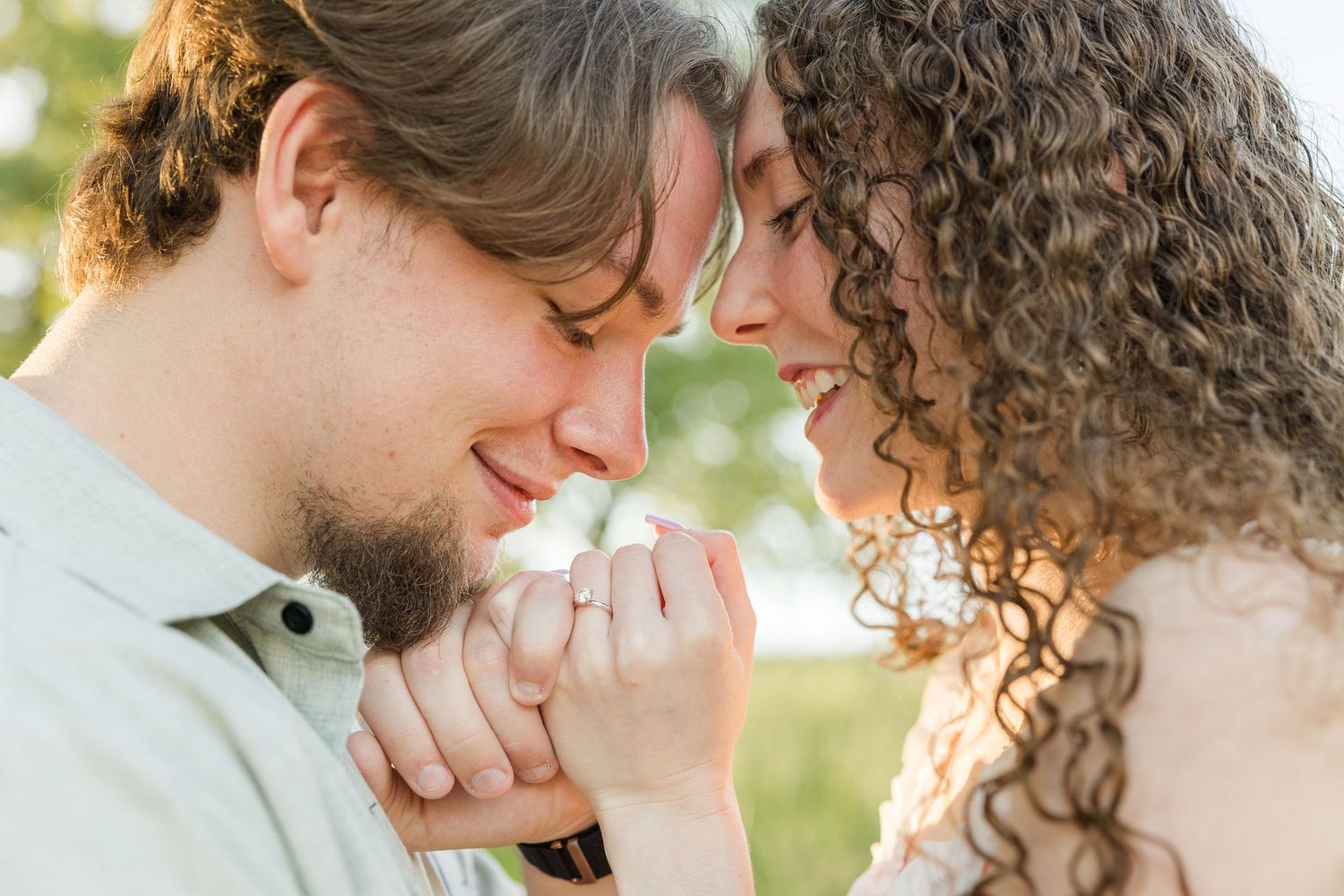 A loving couple shares an intimate moment outdoors, foreheads touching while holding hands in warm sunlight.