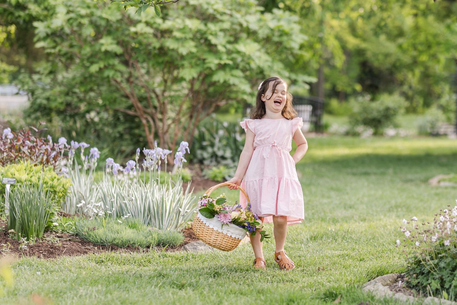 A small child in a pink outfit walks through a lush garden while carrying a wicker basket.