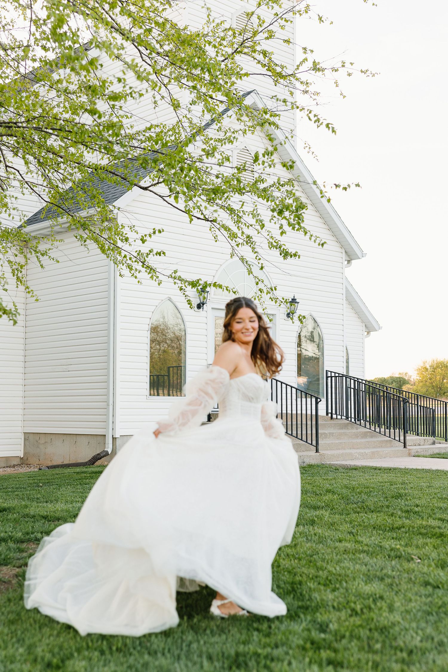 A white wedding dress twirls on a green lawn in front of a white chapel building.