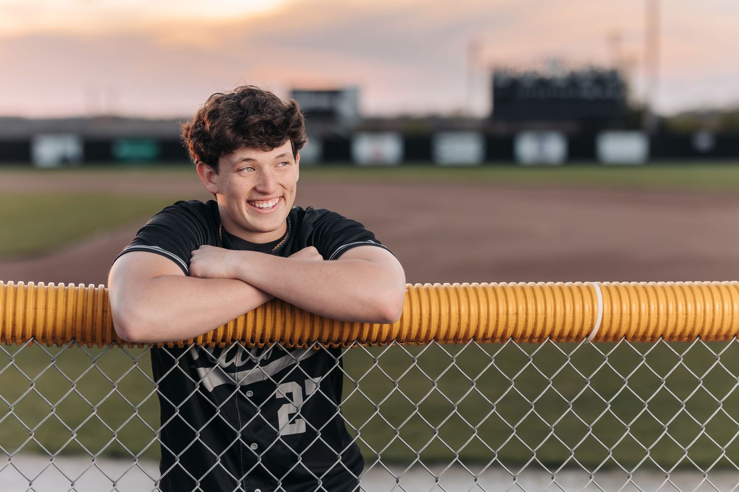 Athletic person in black shirt leaning on yellow fence railing at sports field during sunset.