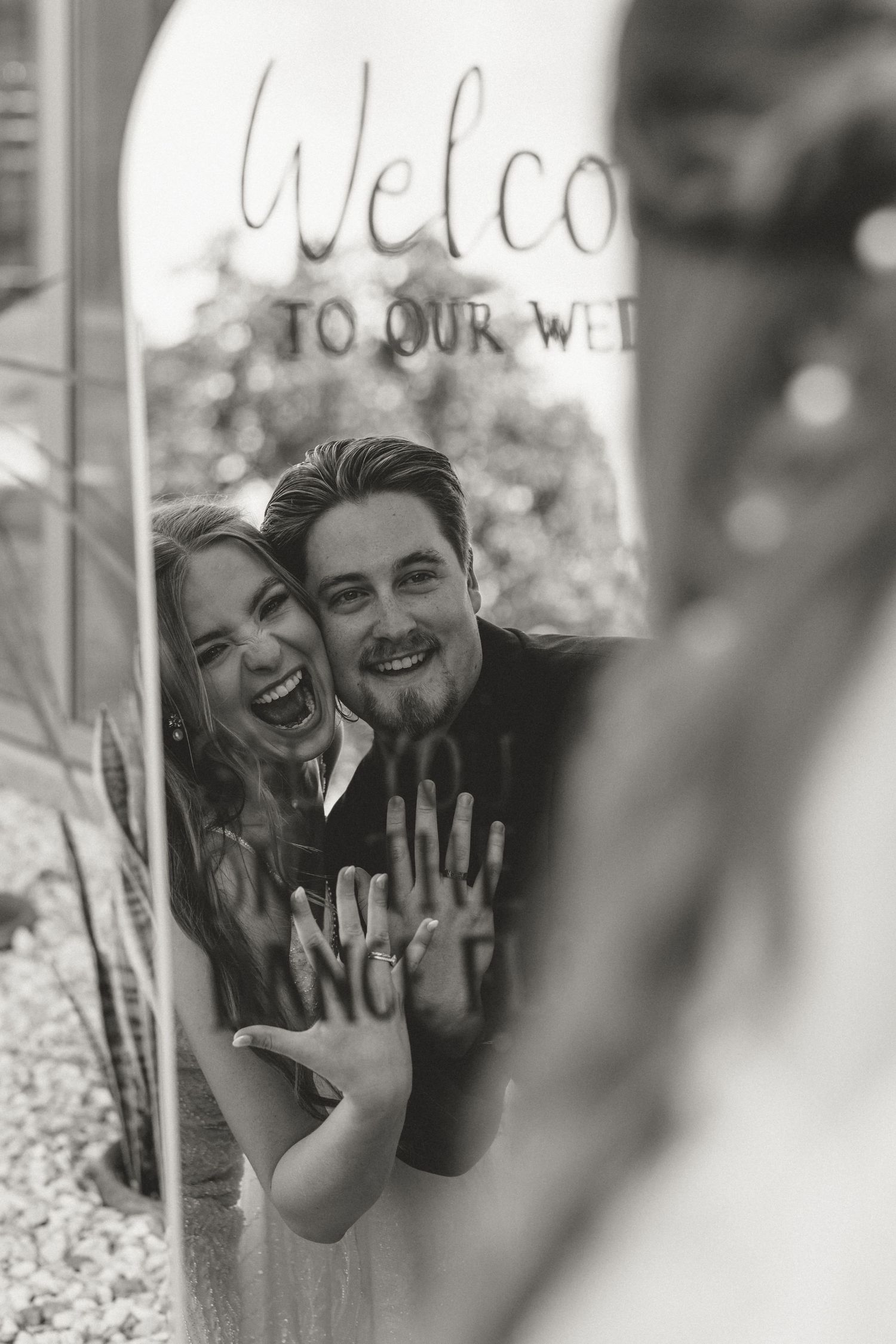 A joyful black and white photo of a couple showing their engagement ring through a glass window.