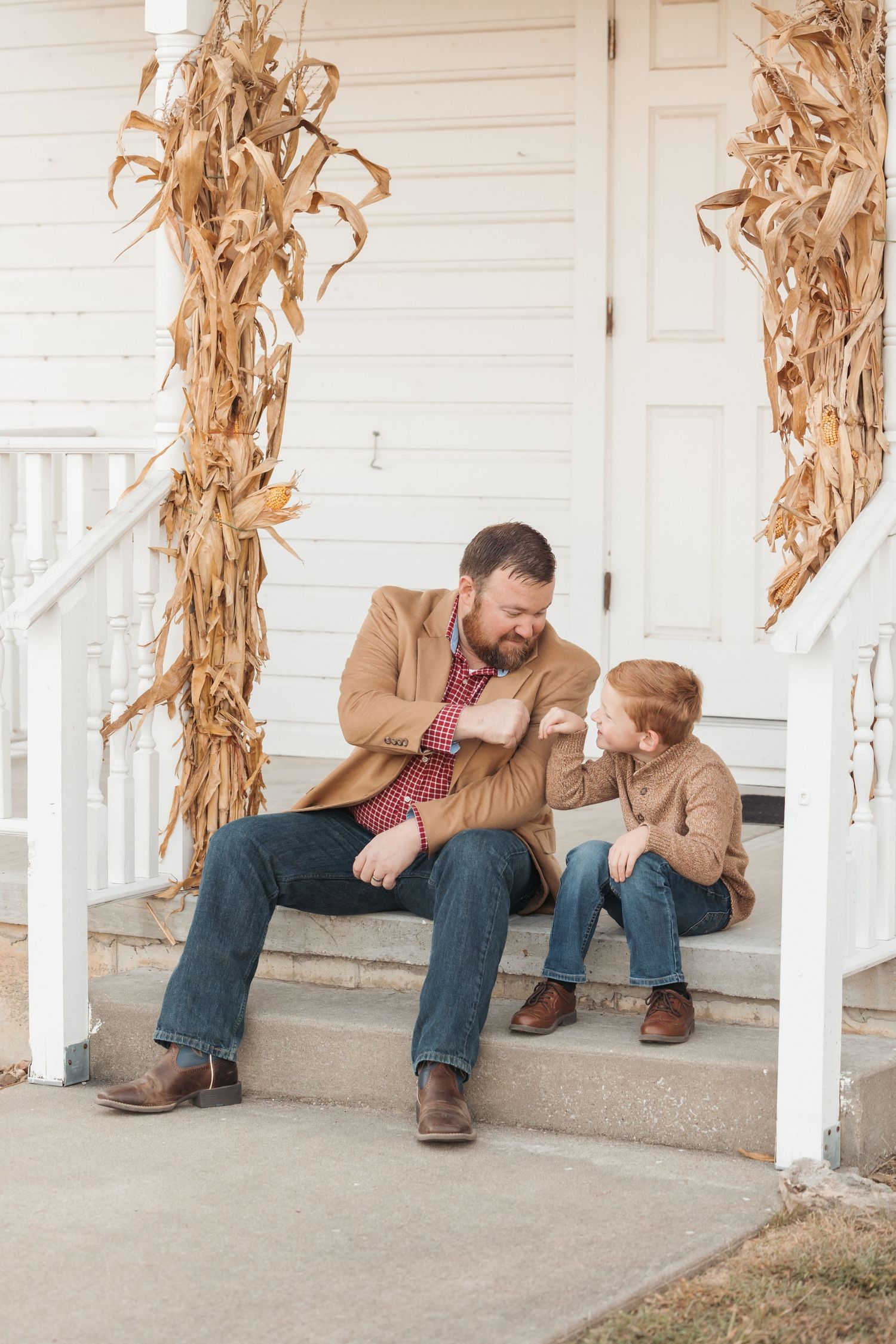 Adult and child sitting on porch steps decorated with dried corn stalks.