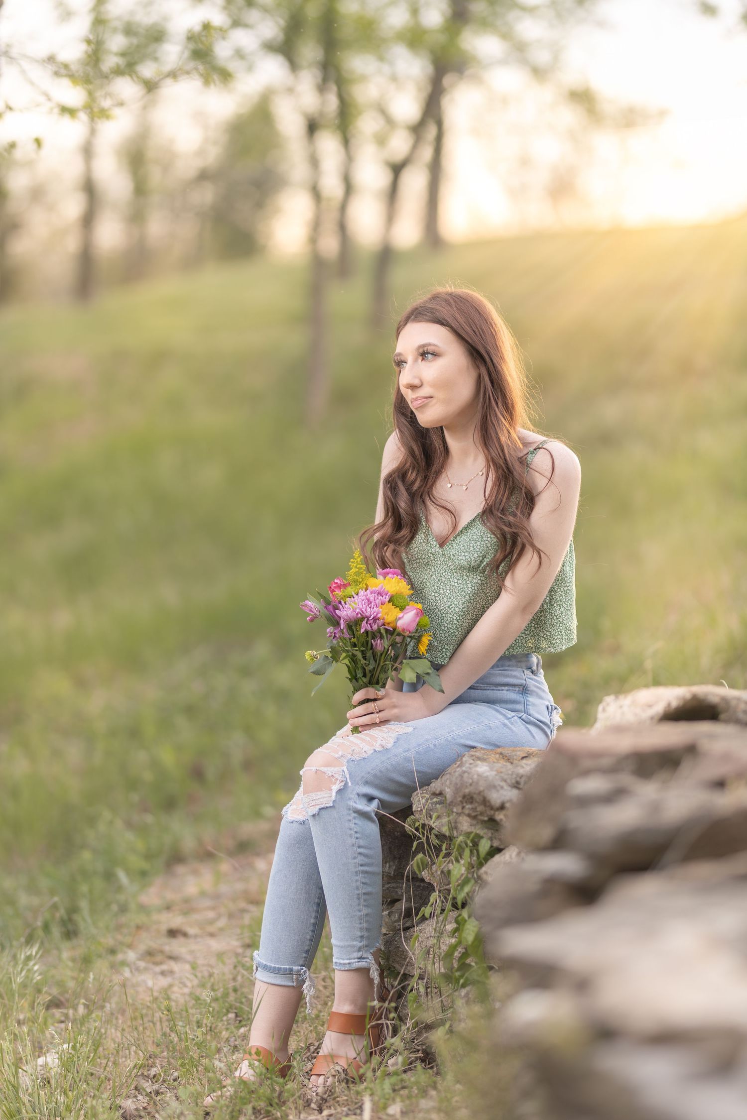 A person sits on rocks at sunset holding colorful wildflowers while wearing casual spring attire.