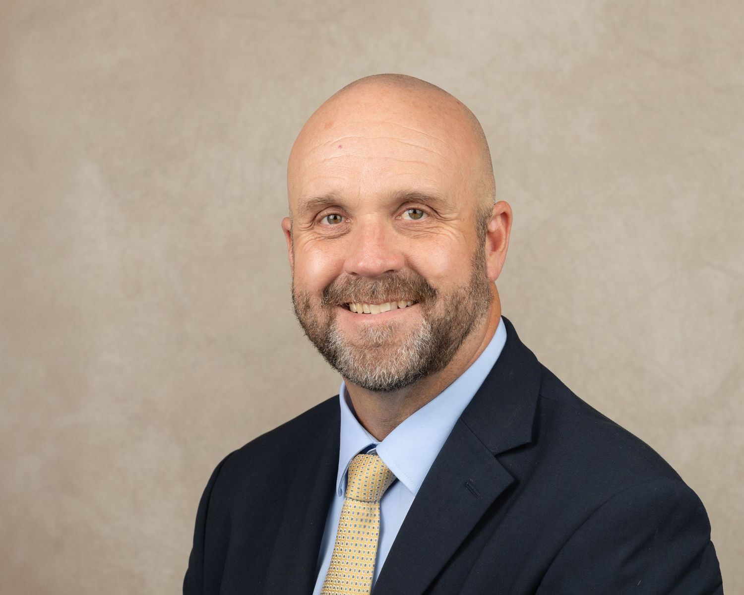 Professional headshot of man in business suit with yellow tie against neutral background.
