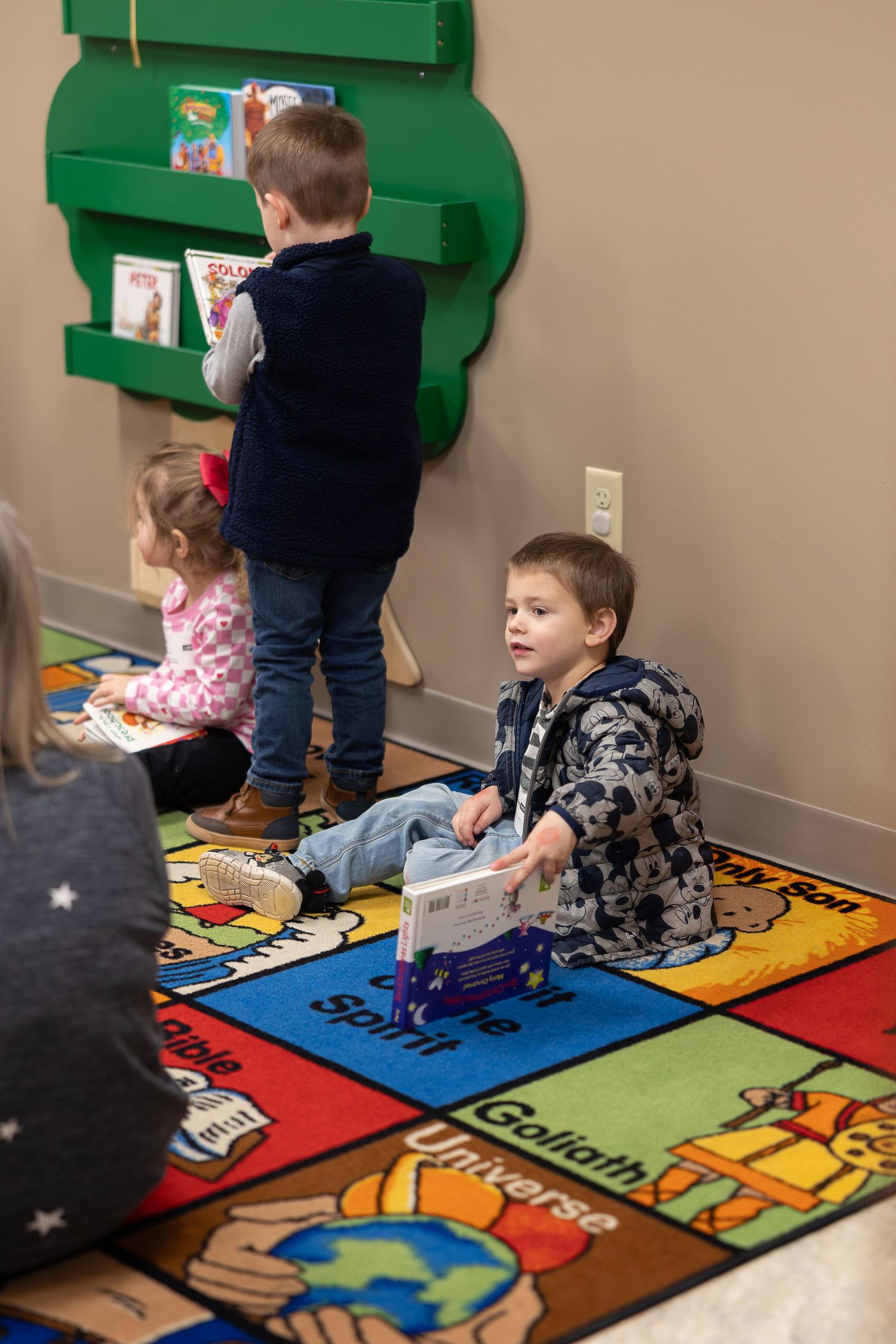 Children explore activities on a colorful educational rug near a green book display.