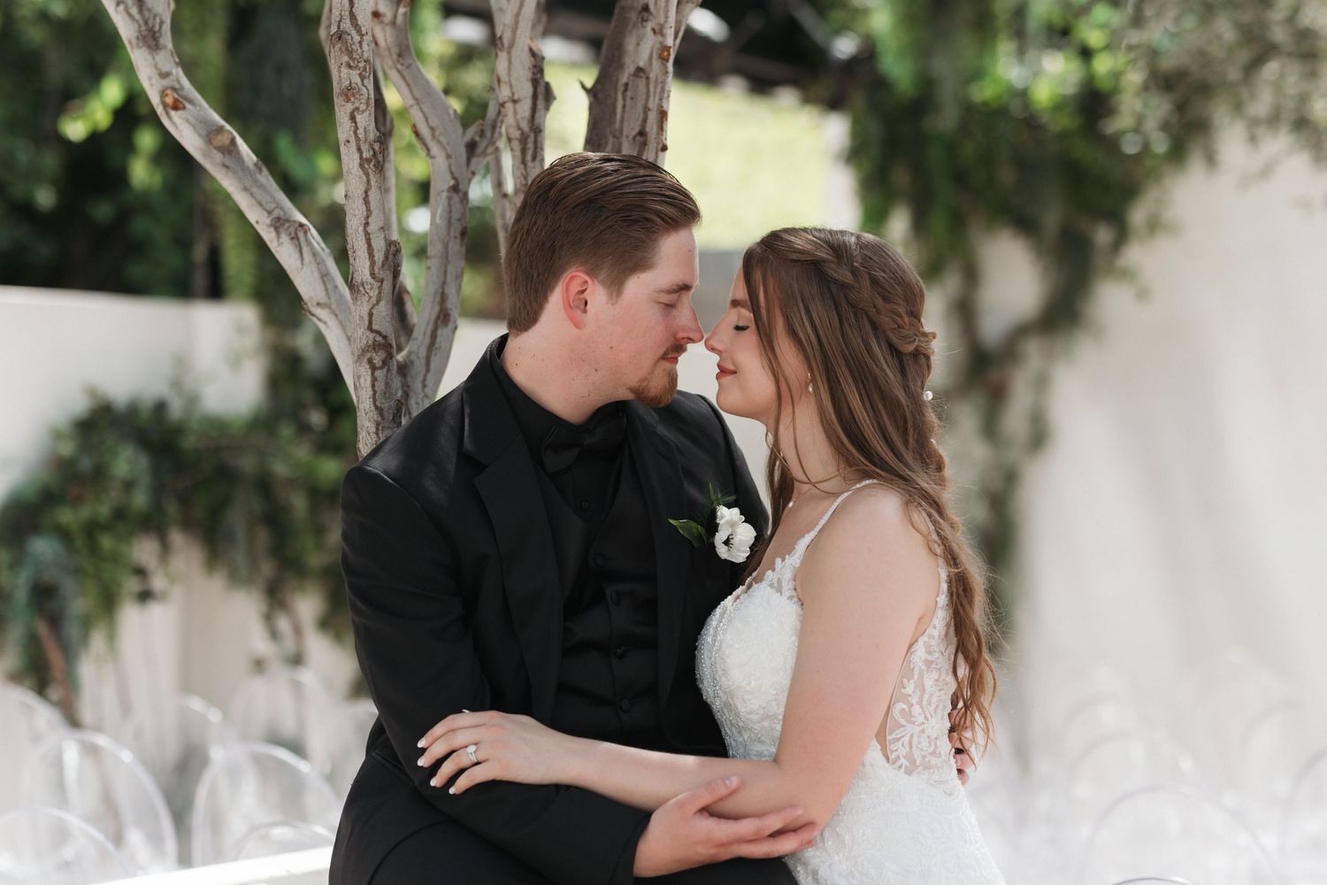 A romantic wedding couple shares an intimate moment together outdoors with white chairs in the background.
