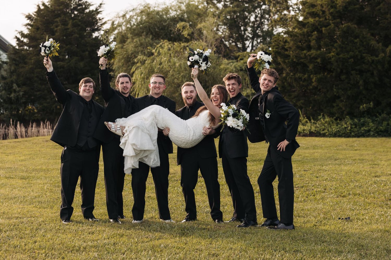 Wedding party members in black attire carry white-dressed figure across outdoor lawn.