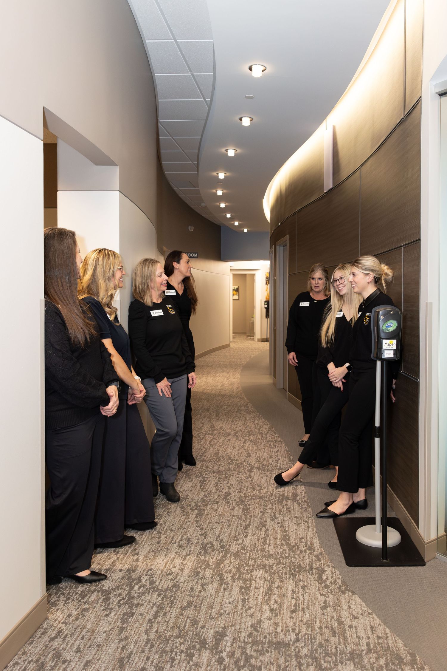 People in black clothing line up along a modern hallway with carpeted floors.