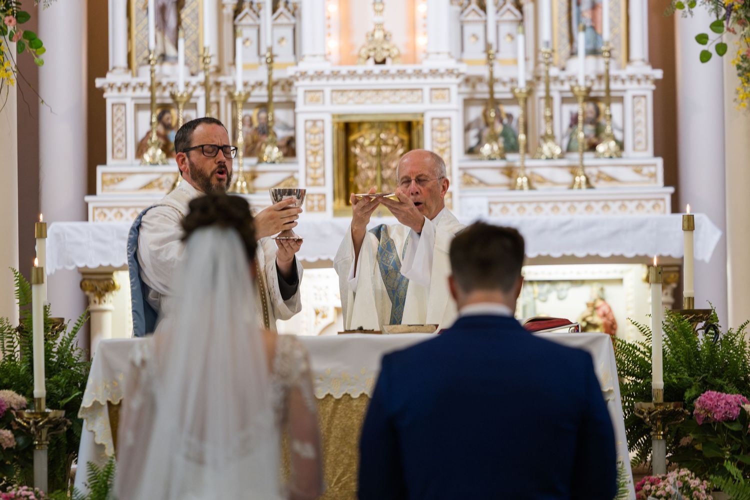 Priest officiating a wedding ceremony at an ornate white altar with floral decorations in a Catholic church.
