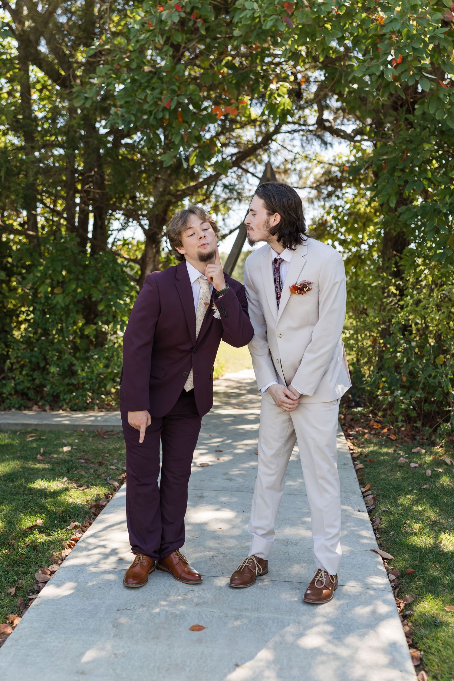 Two young men in suits, one burgundy and one beige, playfully posing on a garden pathway.