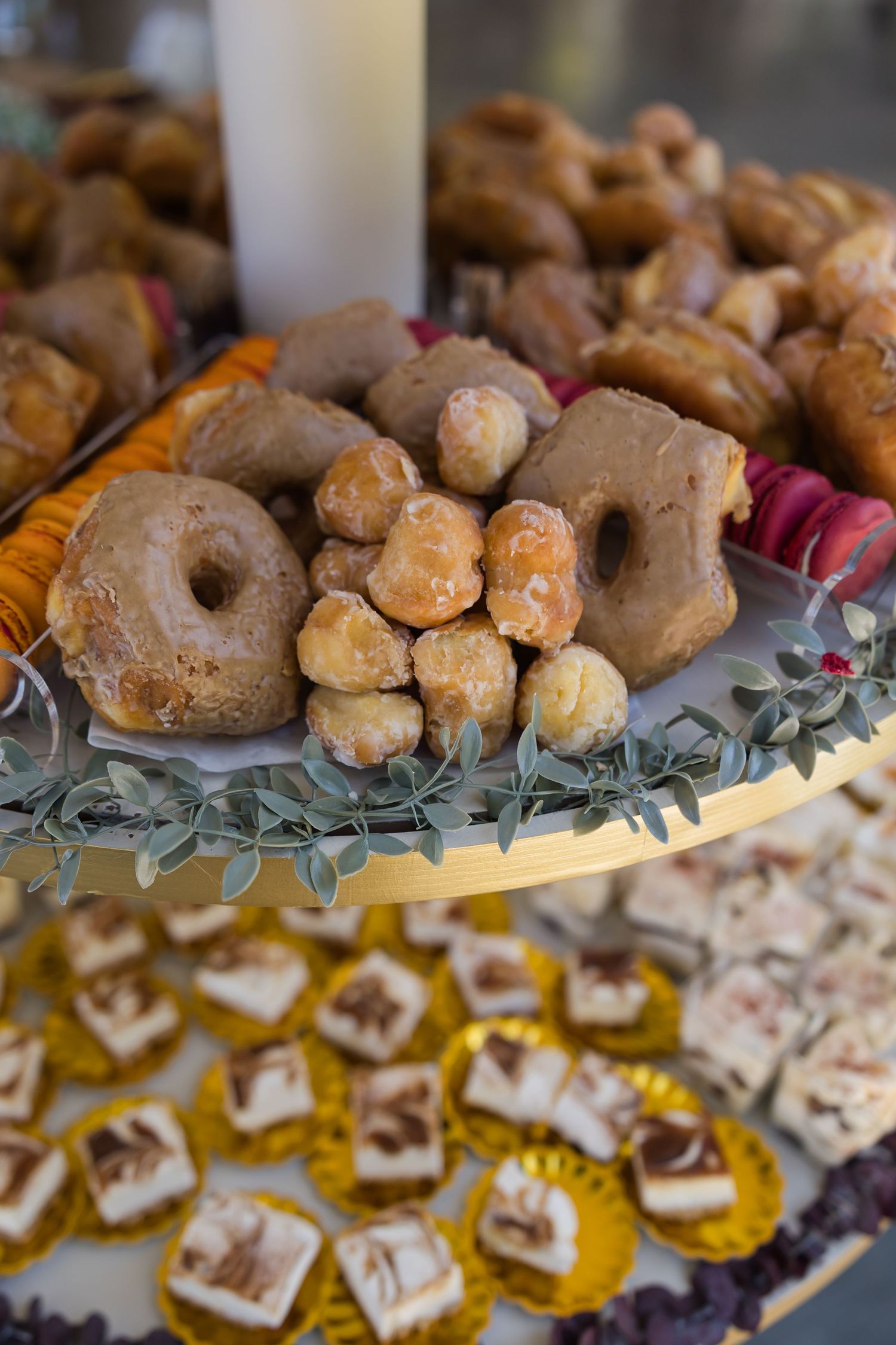Assorted pastries including glazed donuts and donut holes displayed on a tiered dessert stand with macarons and fudge.