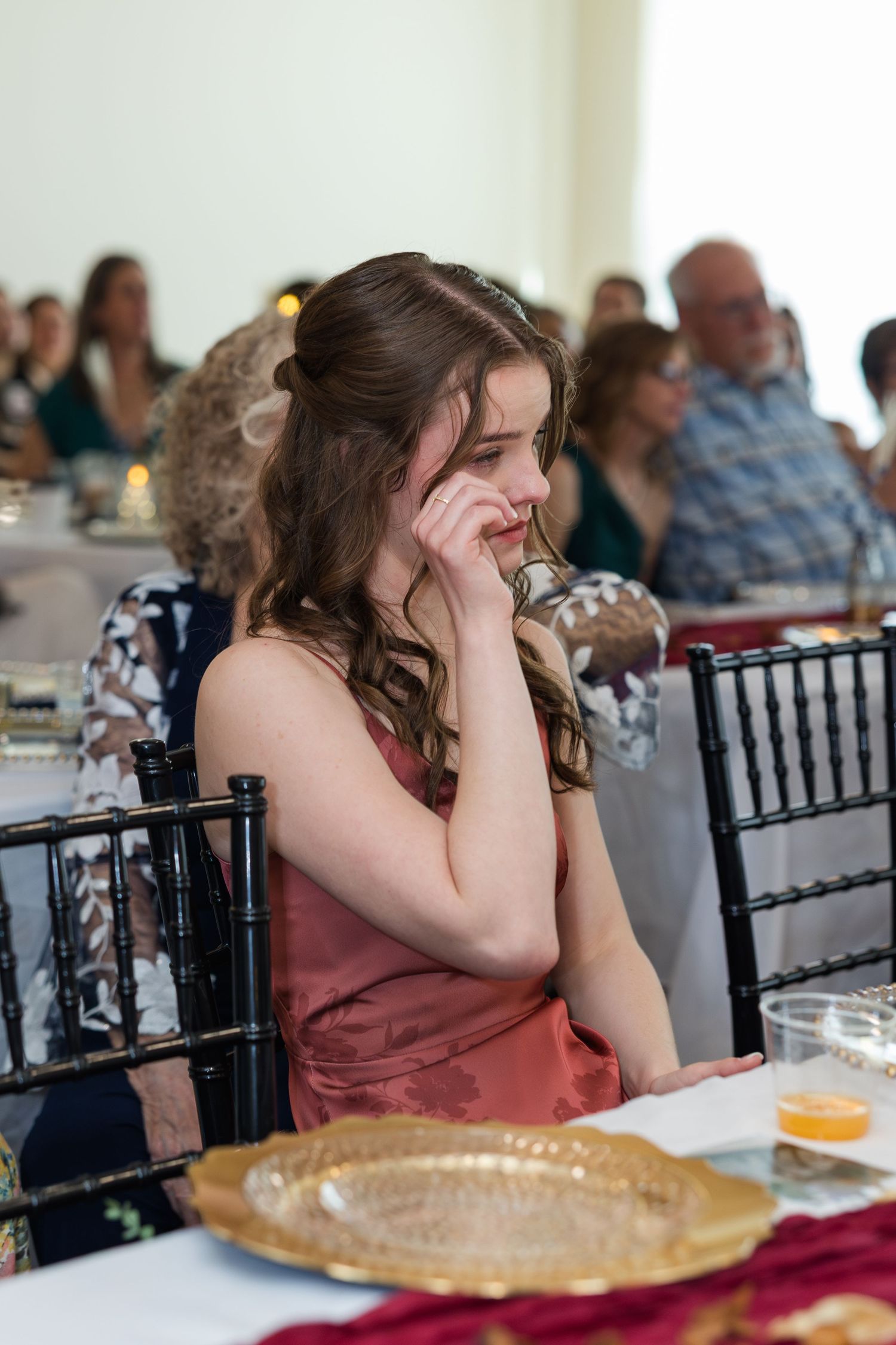 Young bridesmaid in coral dress sits pensively at wedding reception table with gold charger plate.
