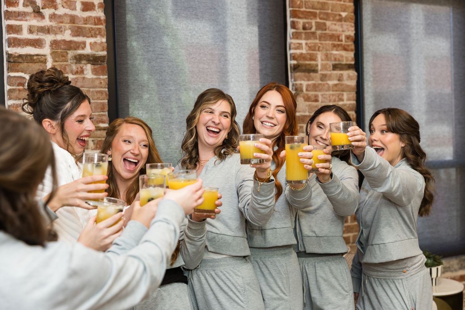 Bridesmaids in grey robes toasting with orange juice glasses, laughing and celebrating together indoors.