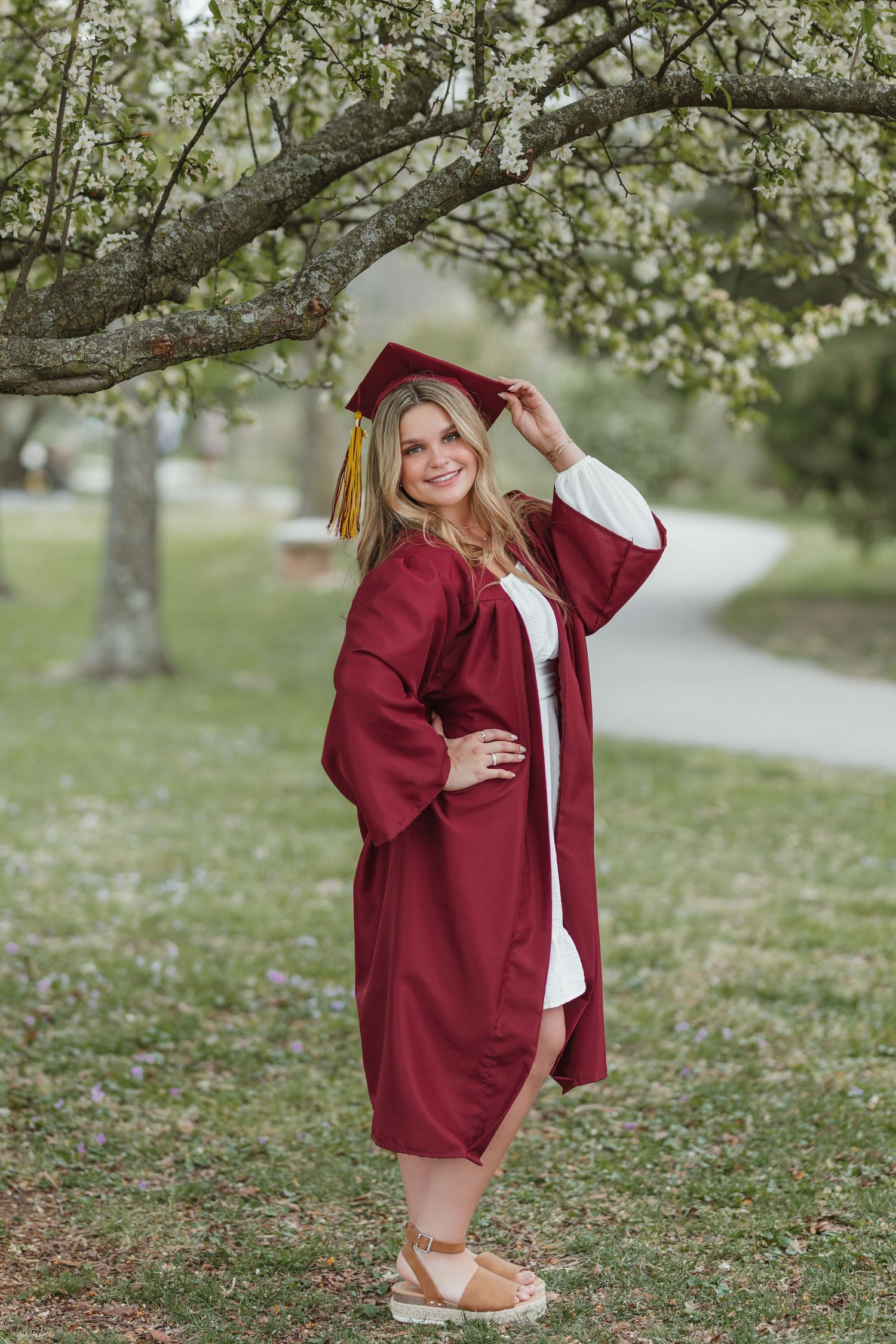 Graduate in maroon cap and gown poses under blooming spring tree during outdoor graduation photo session.