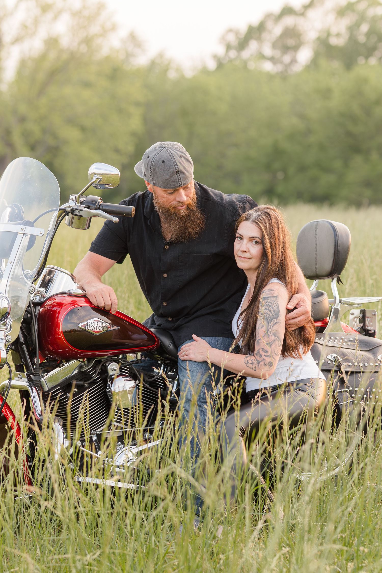 A couple sits together on a Harley Davidson motorcycle parked in a tall grass meadow during a summer photoshoot.