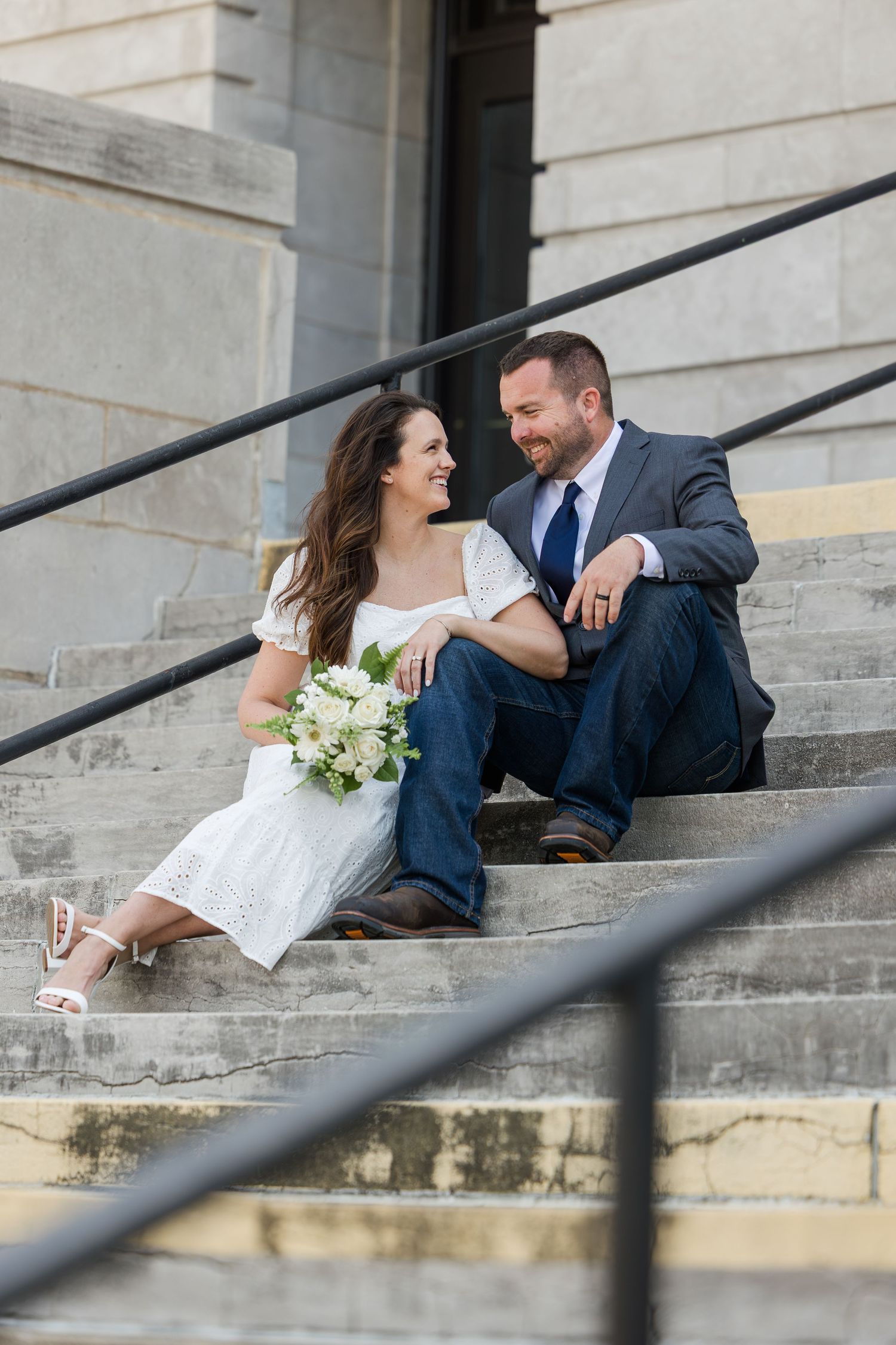 A couple wearing formal attire sits on stone steps together outside a grand building.