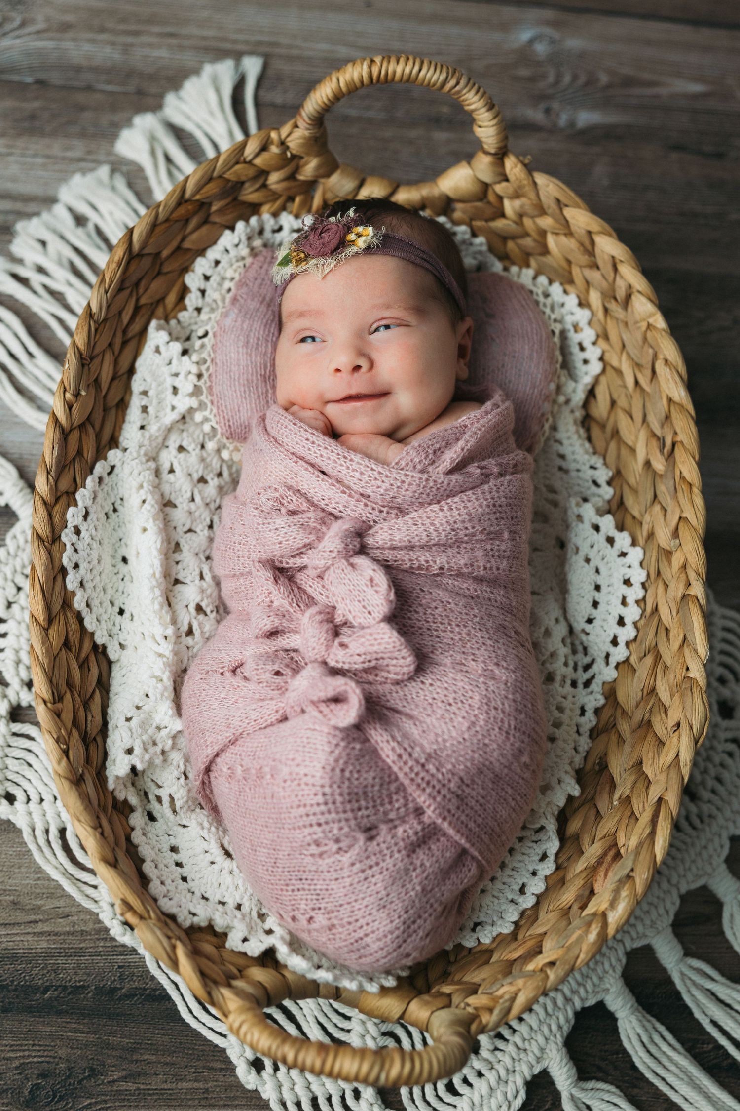A newborn wrapped in pink knit fabric rests on a white lace blanket inside a woven basket.