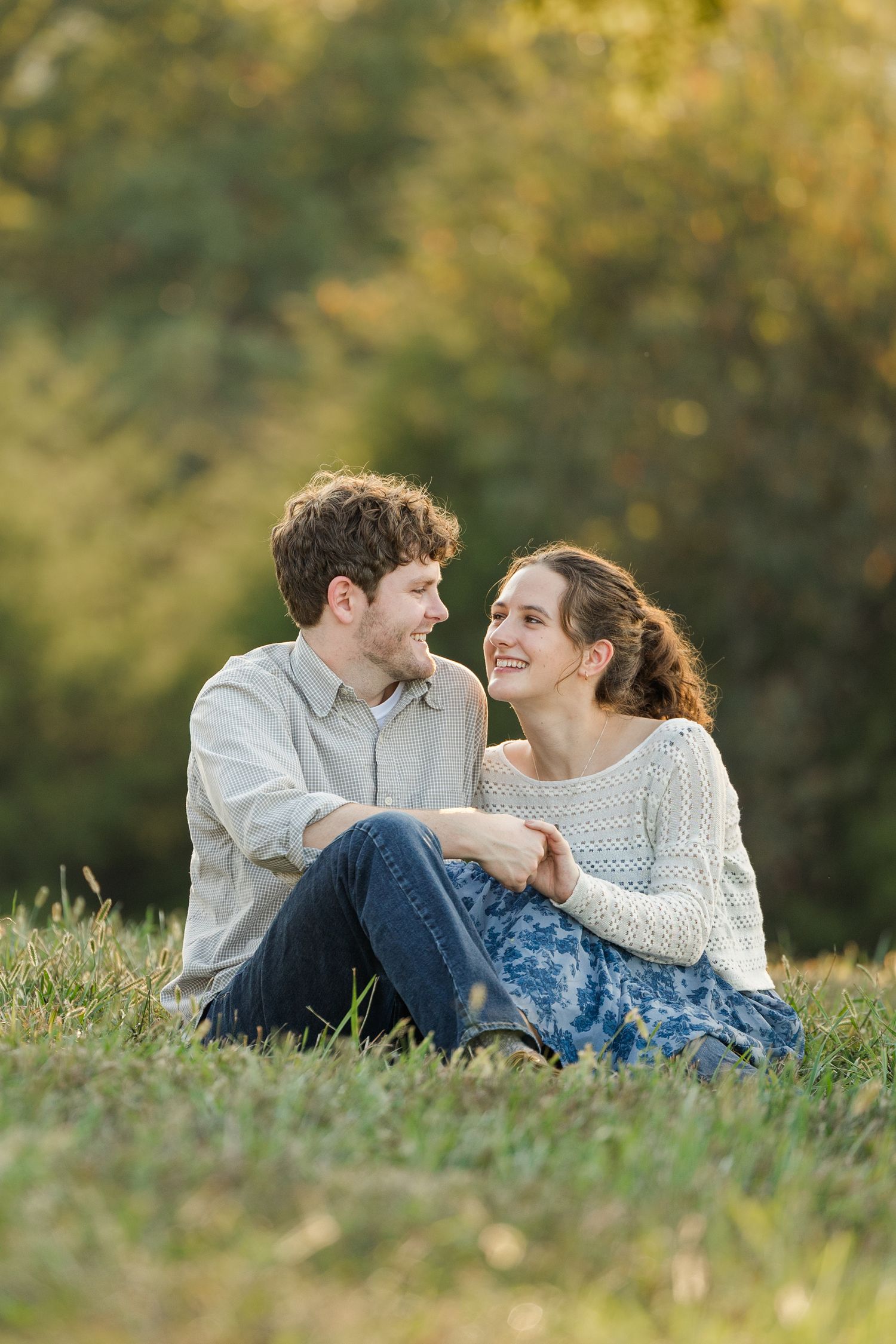 A romantic couple sits together on grass during golden hour, wearing casual sweaters and sharing an intimate moment.