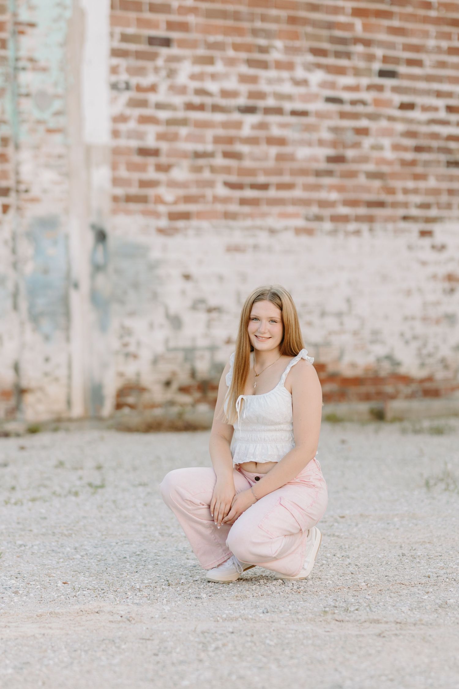 A figure in a white crop top and pink pants poses casually against a brick wall outdoors.