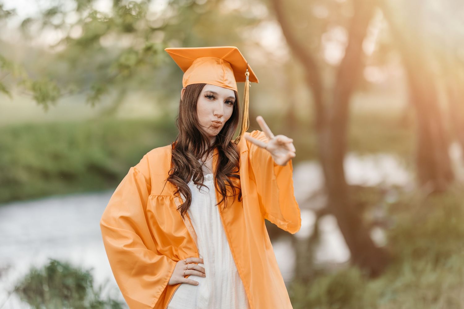 Graduate in orange cap and gown poses outdoors in natural setting with sunlit trees in background.