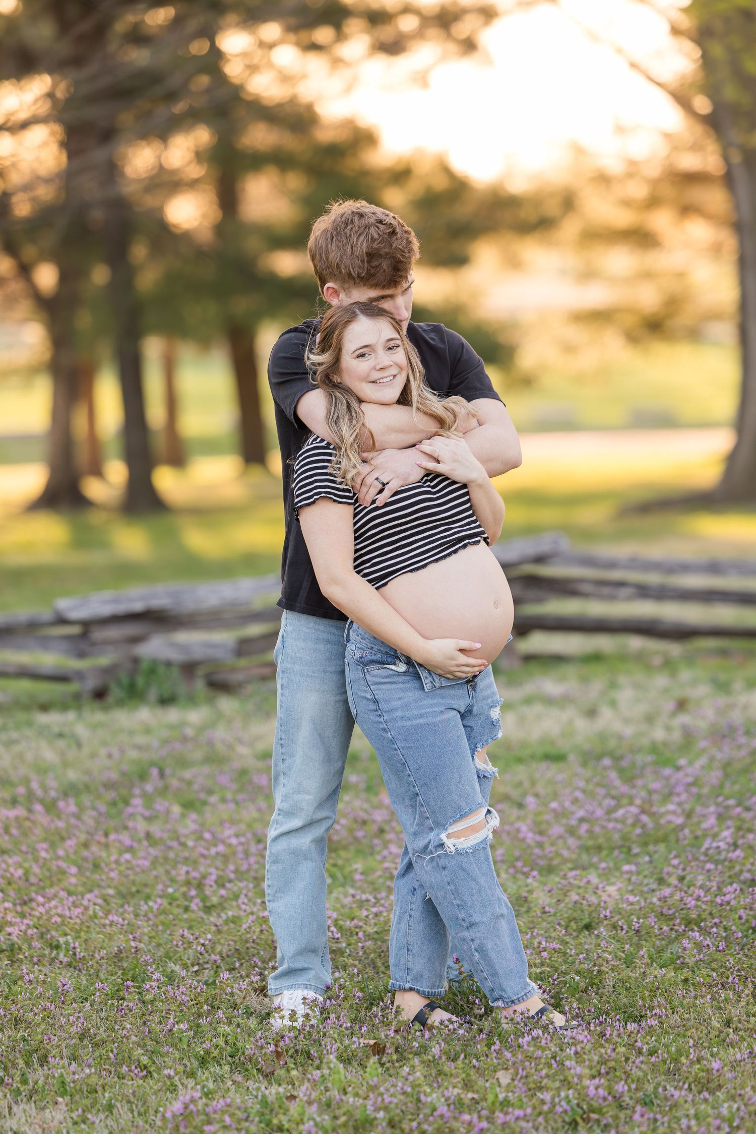 A maternity photo of a couple embracing outdoors at sunset in a park setting.