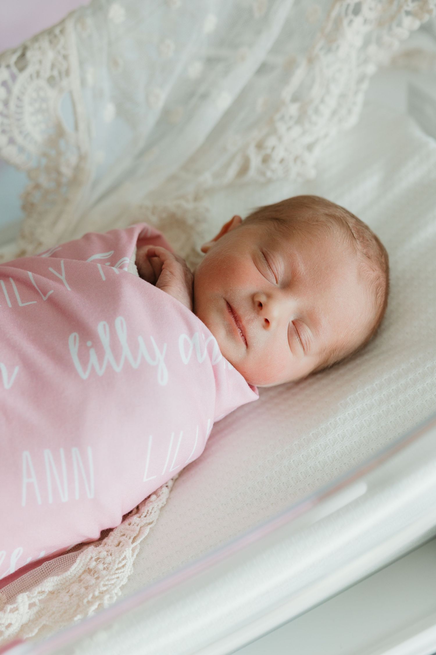 Sleeping newborn baby wearing pink swaddle wrap rests peacefully on white lace bedding.