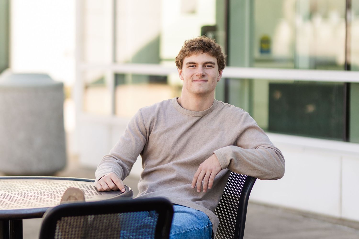 Casual portrait in beige sweater while sitting at outdoor table with modern building exterior.