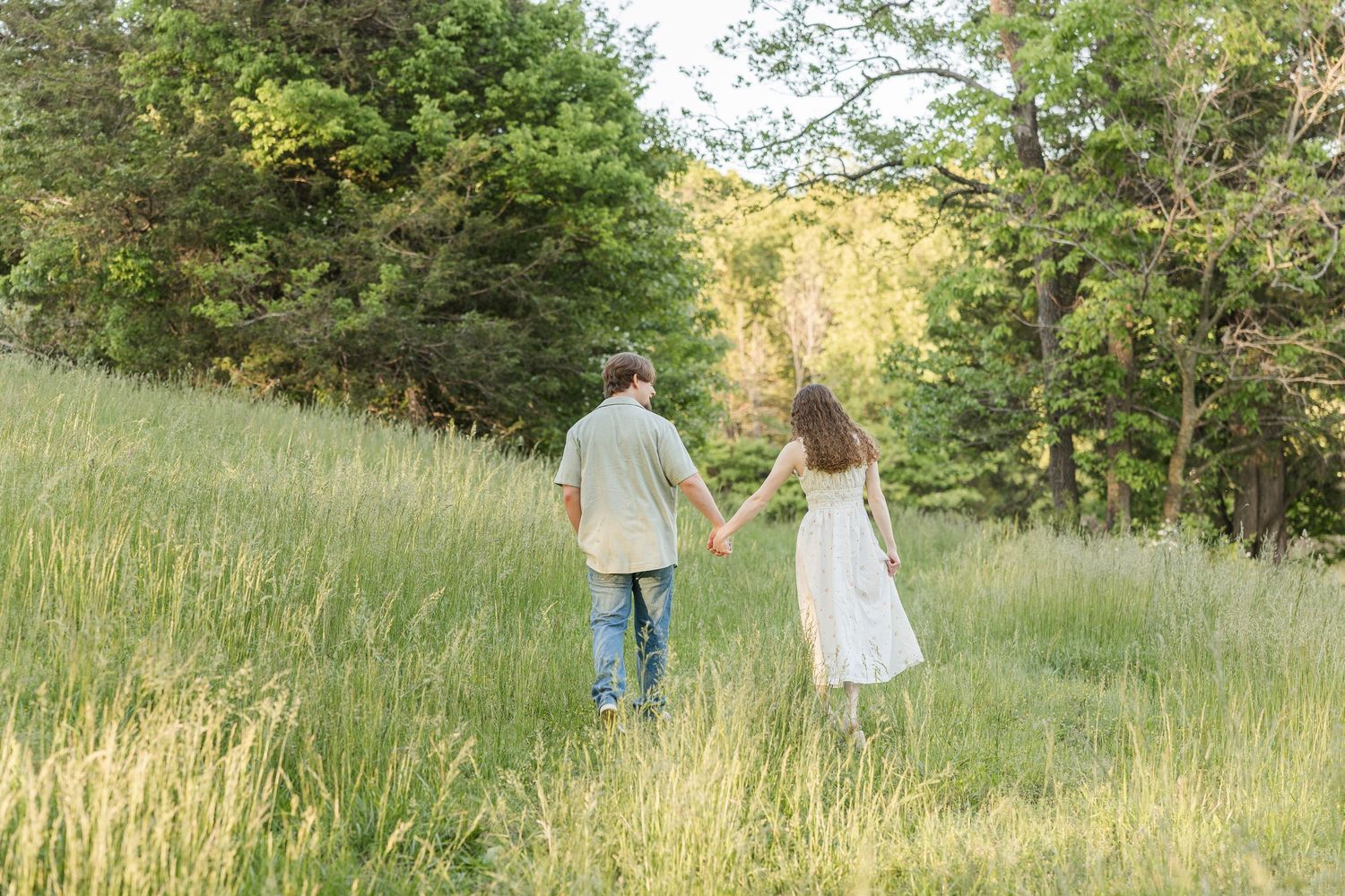 Couple walking hand in hand through tall grass on a grassy hillside surrounded by trees.