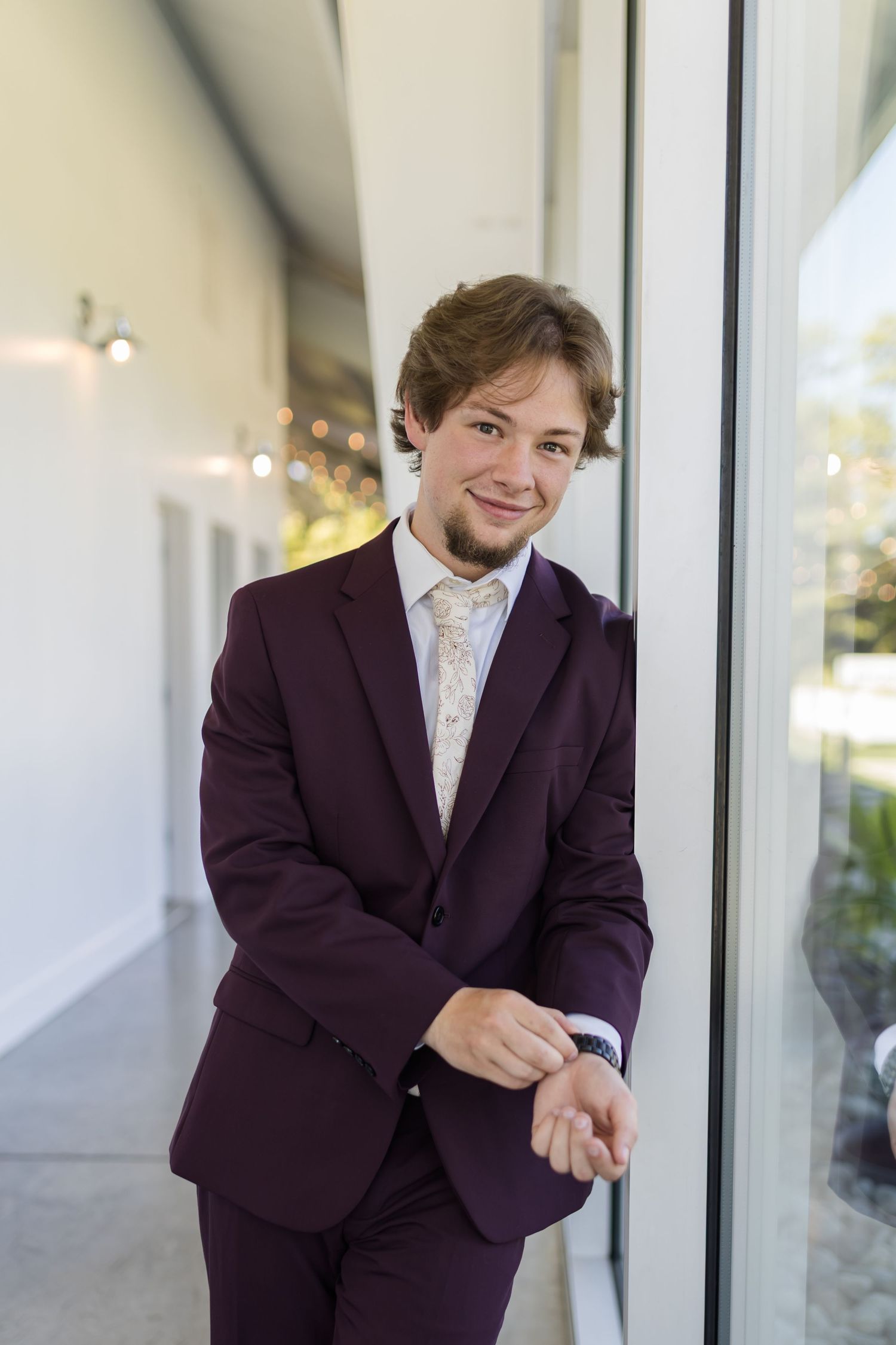 Young groom in burgundy suit and floral tie leans against window smiling at wedding venue.