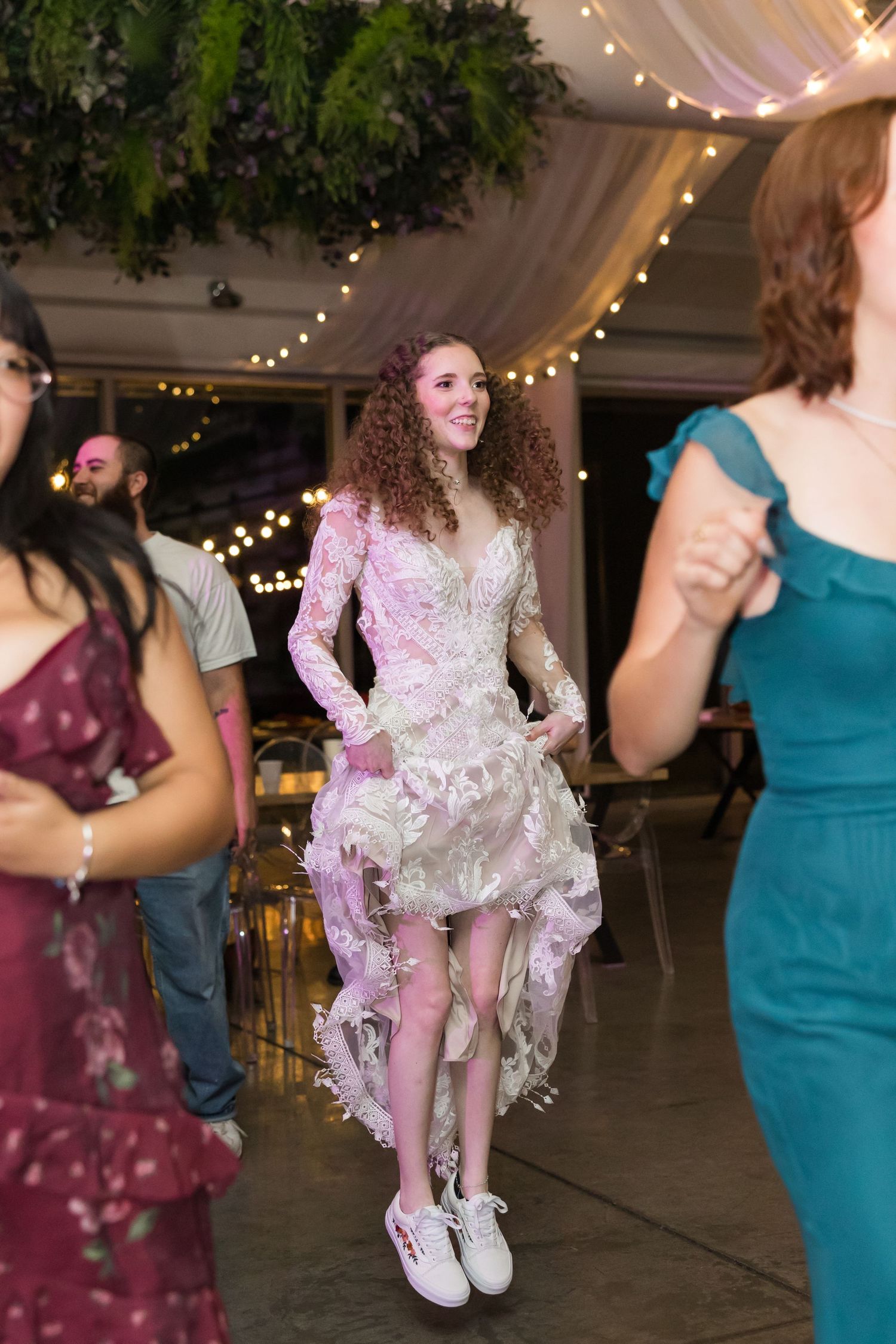 Bride in lace hi-lo dress and sneakers dances joyfully with guests at wedding reception.