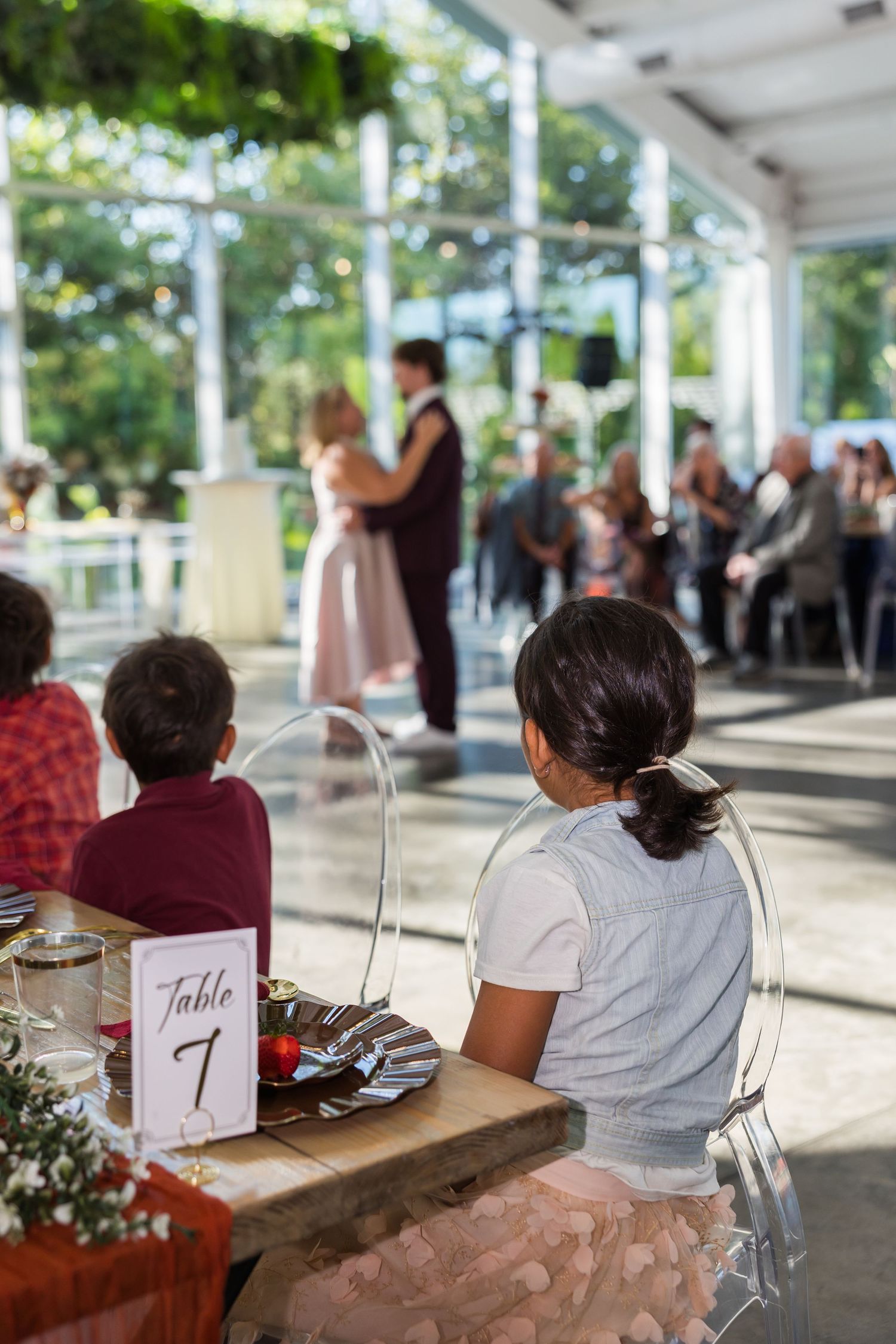 Two children sit at Table 7 watching couple dance on floor of sunlit greenhouse wedding reception.