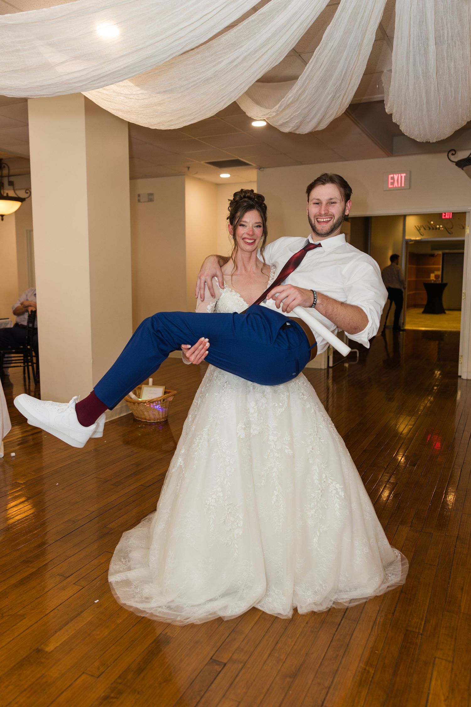 Laughing groom carries bride in wedding dress on dance floor during lively reception celebration.