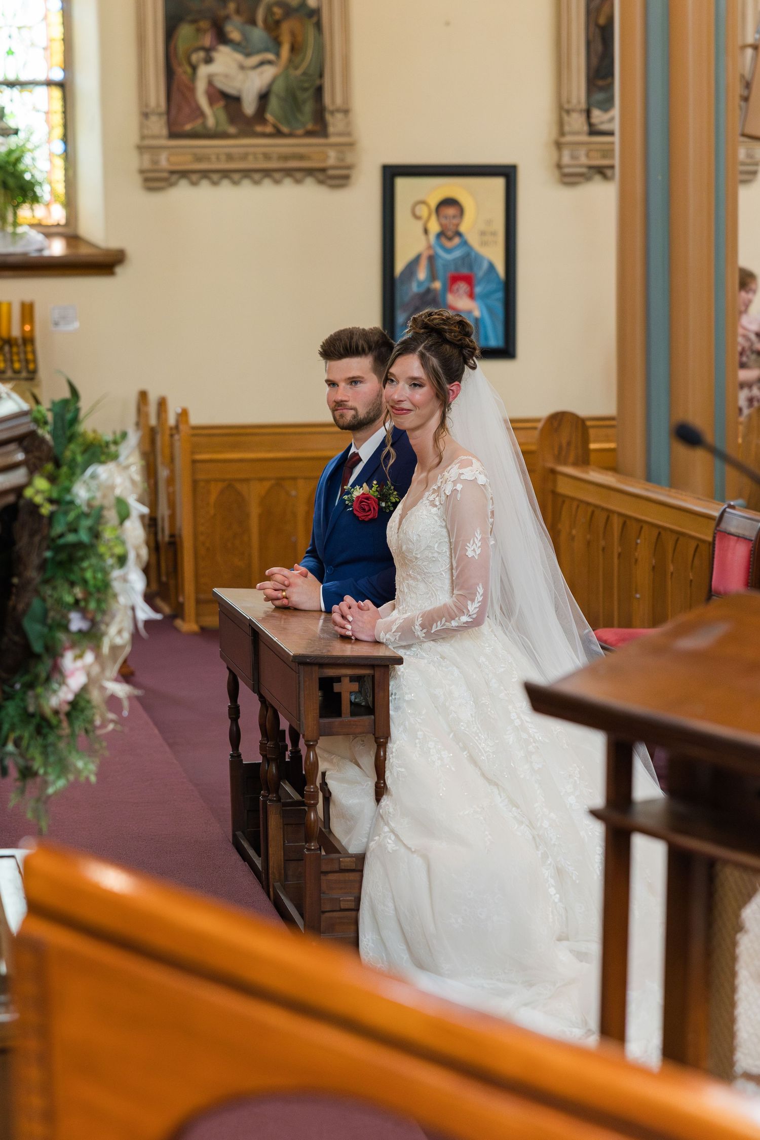 Bride and groom kneel together at a wooden prie-dieu during their Catholic church wedding ceremony.