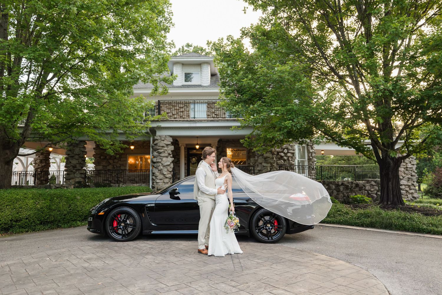 Bride and groom pose beside a black Porsche sports car in front of an elegant stone venue surrounded by trees.
