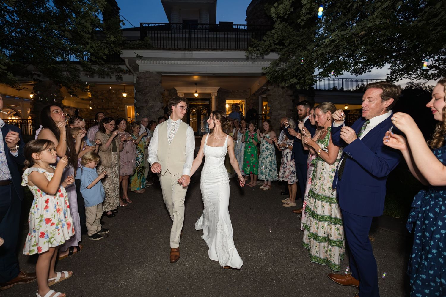 Bride and groom hold hands walking through cheering guests at outdoor evening wedding reception venue.