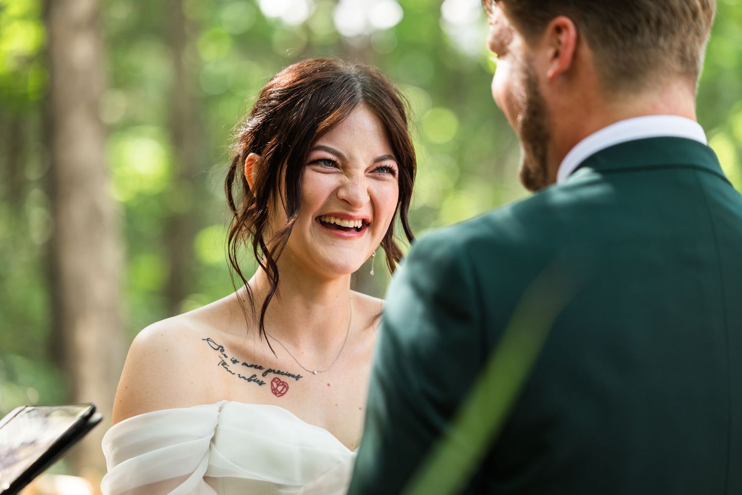 Bride laughing joyfully during outdoor wedding ceremony with groom in green suit amid forest backdrop.