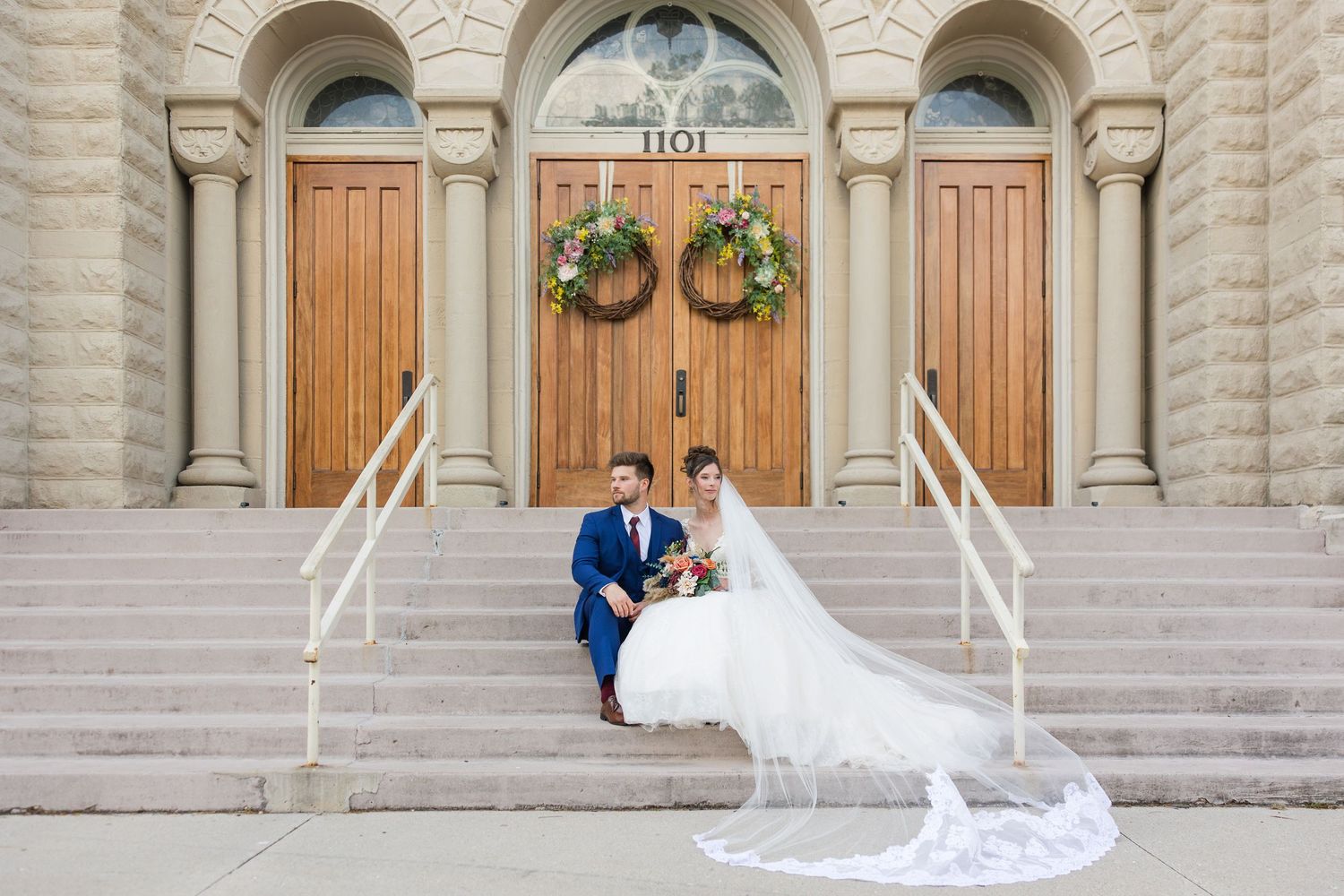 Bride and groom sitting on church steps in wedding attire, long veil flowing down stairs.