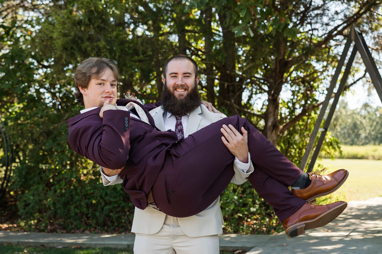 Bearded groom in tan suit carries groomsman in burgundy suit outdoors at wedding celebration.