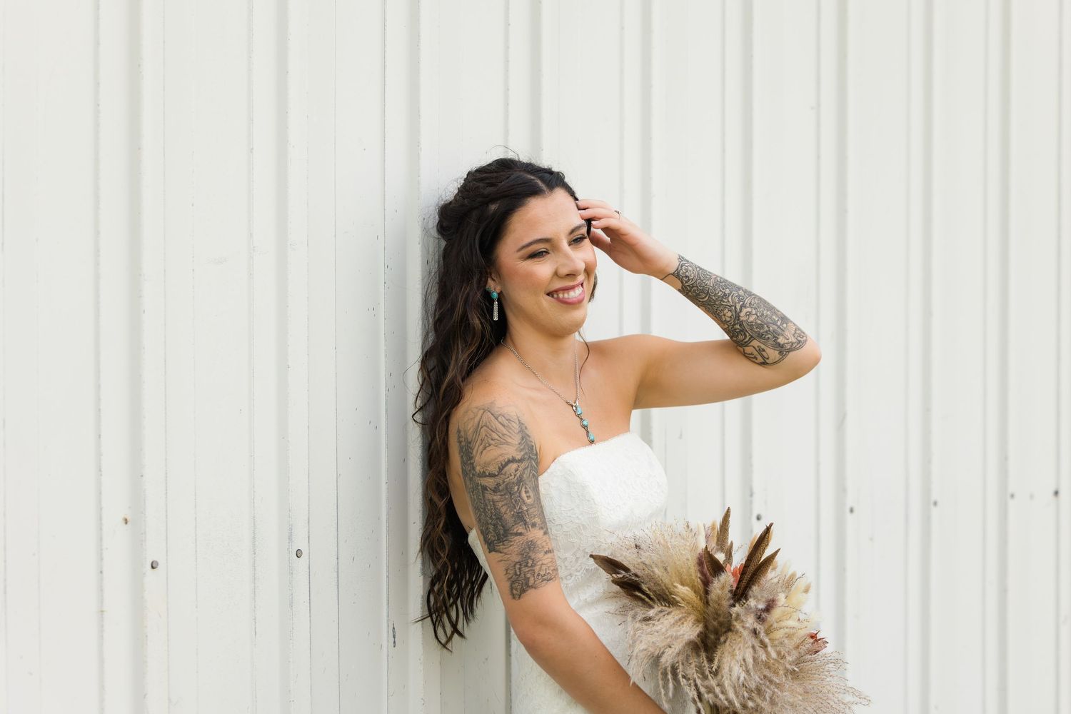 Smiling bride in white strapless gown with tattoos holding a dried floral bouquet against a white wall.