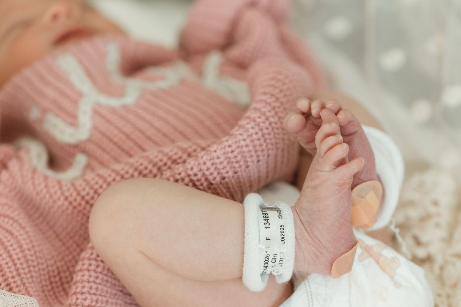 A newborn baby wears hospital ID bracelets while wrapped in a soft pink knit blanket.