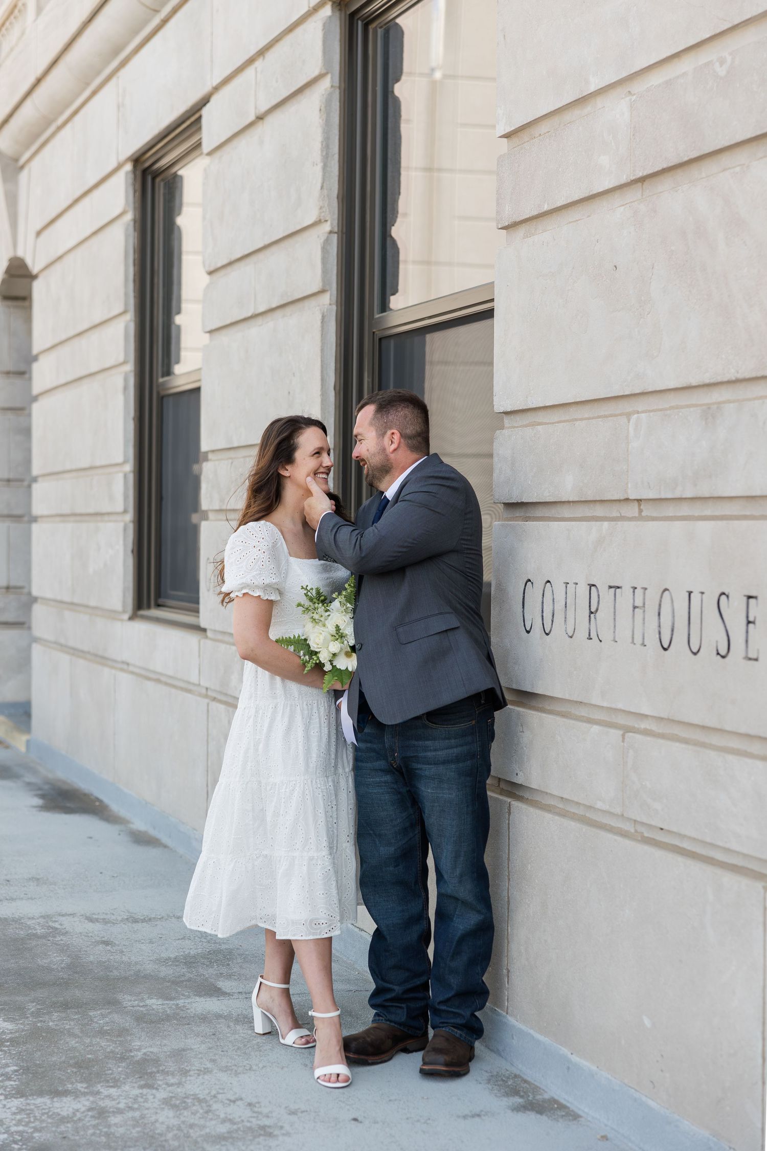 A couple stands intimately against a courthouse wall during their wedding ceremony, with the bride holding a white bouquet.