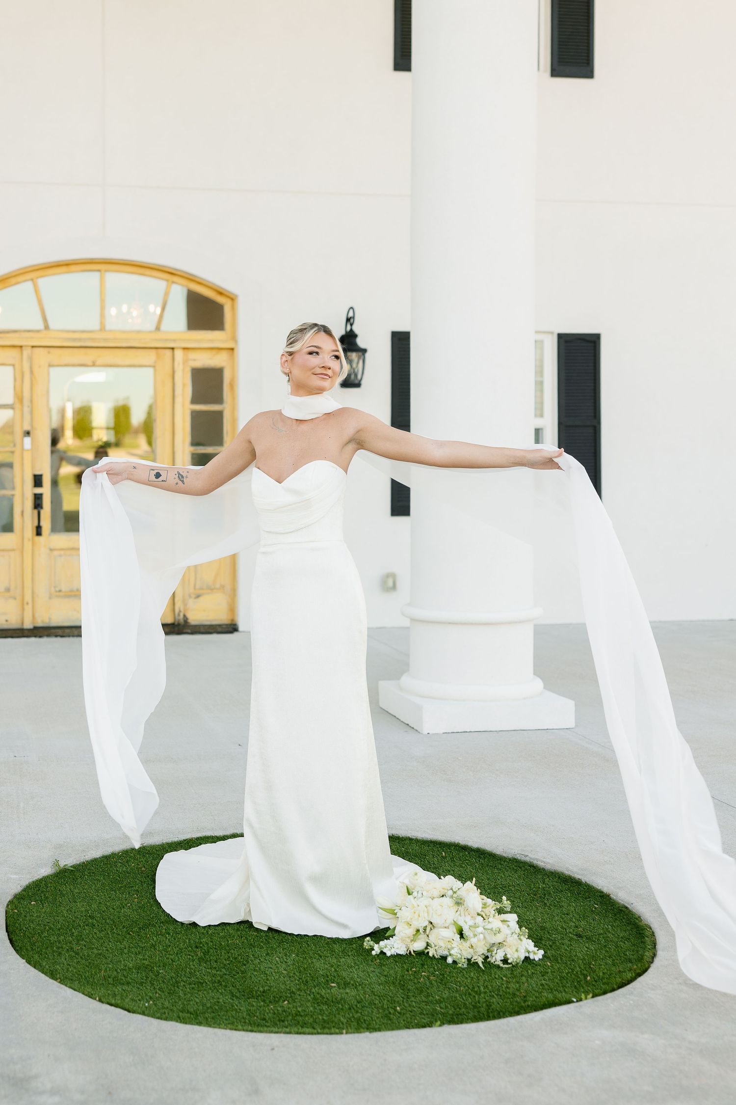Elegant bride in sleek white column gown with flowing cape sleeves poses on circular grass patch with white bouquet.