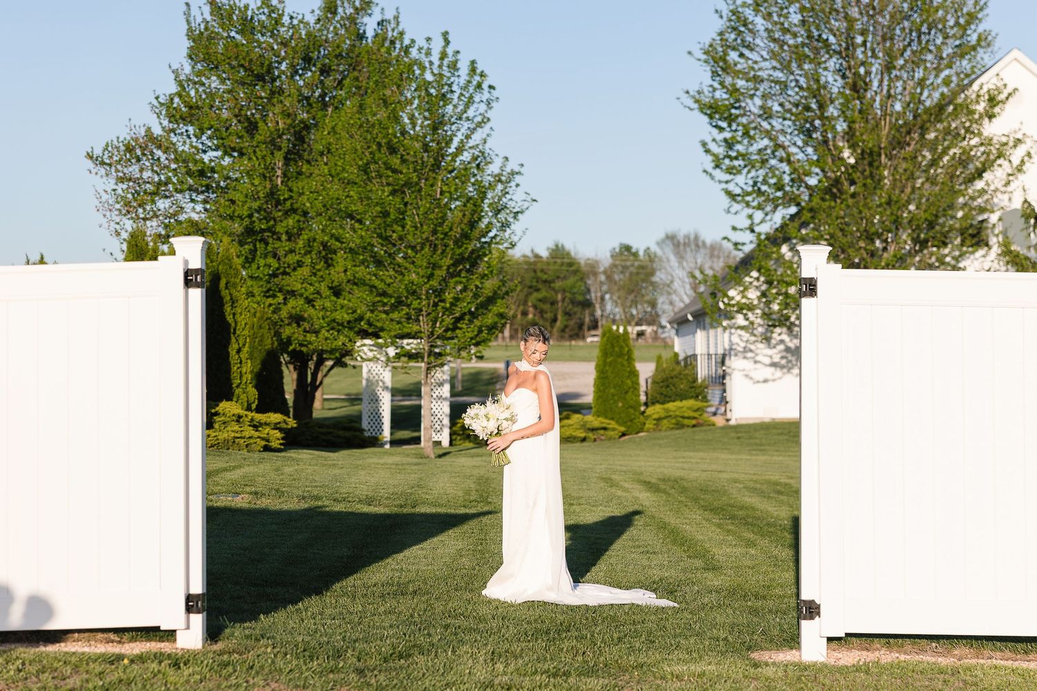 A wedding ceremony setup outdoors with white partitions, green trees and a grassy lawn on a sunny day.