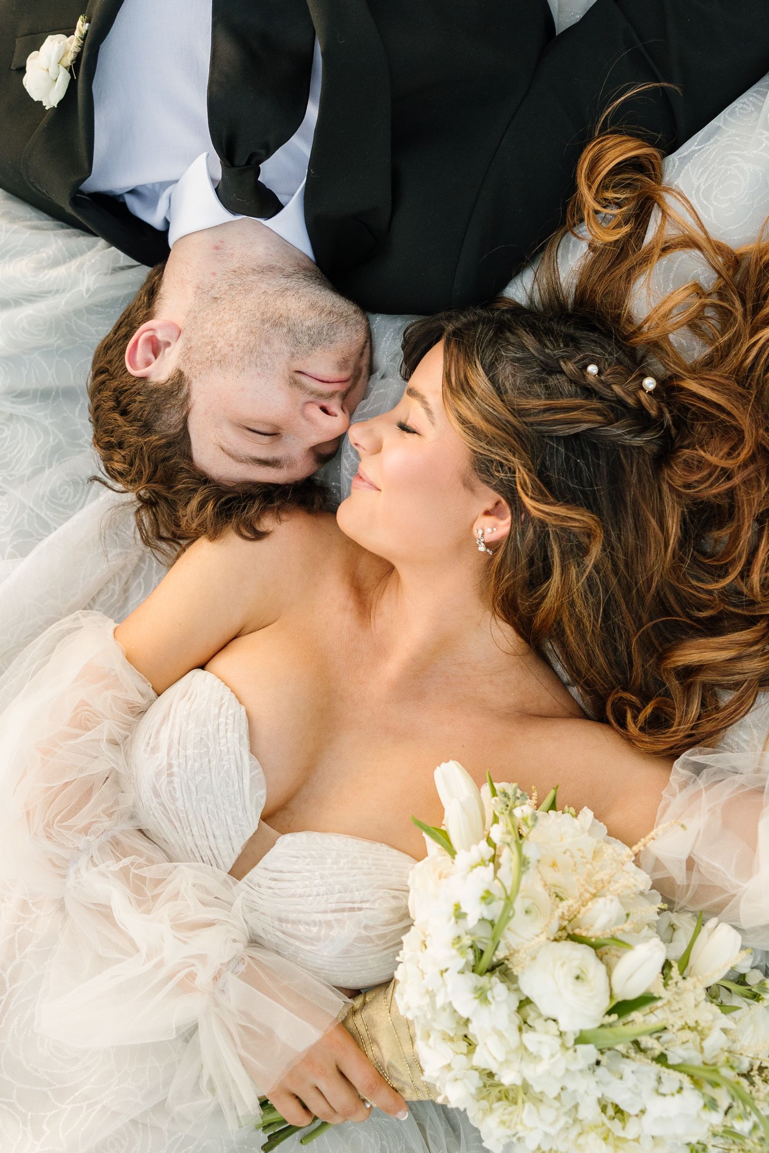 Bride and groom lying down together, bride holding white floral bouquet, sharing an intimate moment in wedding attire.