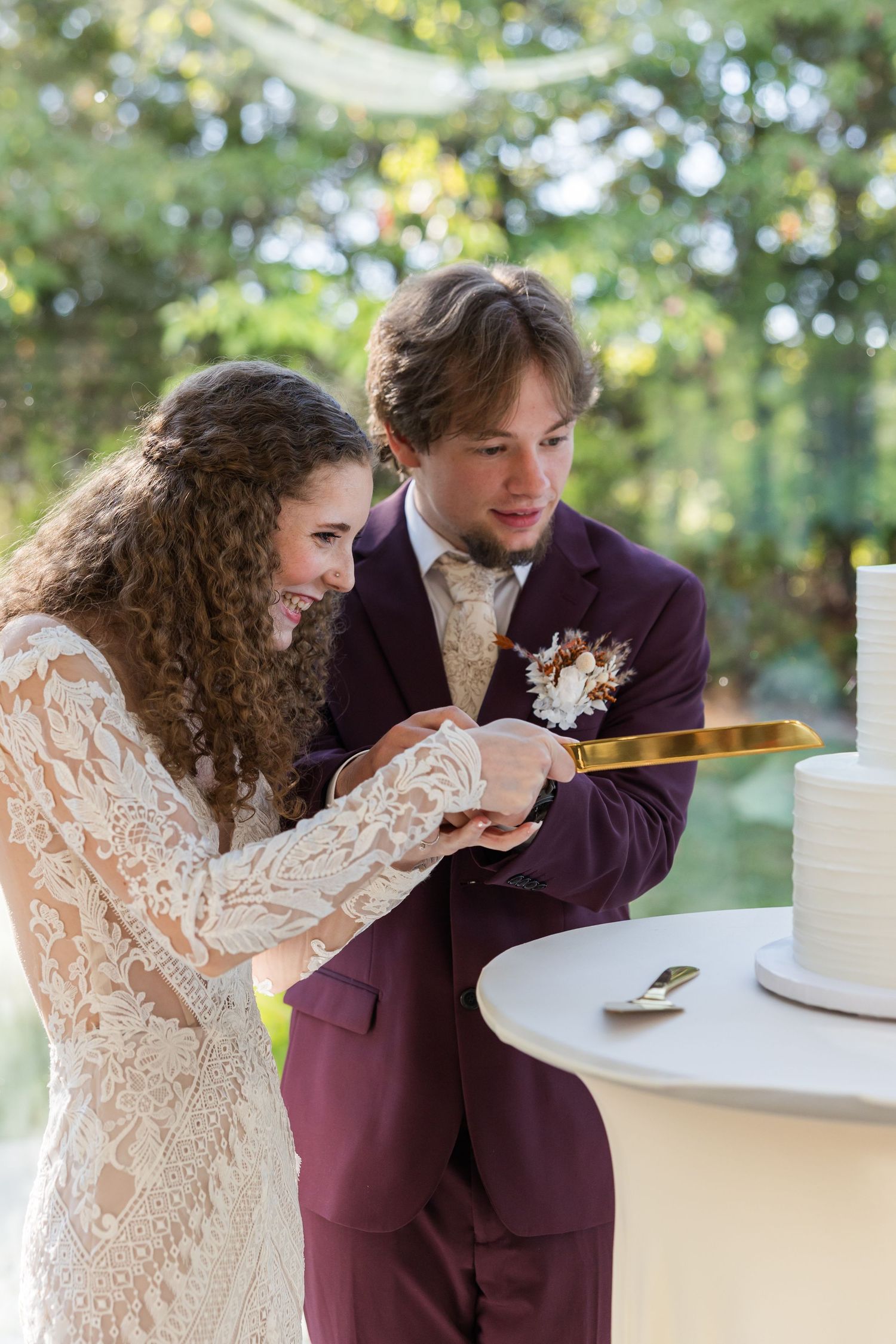 Bride and groom cutting their wedding cake outdoors, she in lace gown, he in burgundy suit with boutonniere.