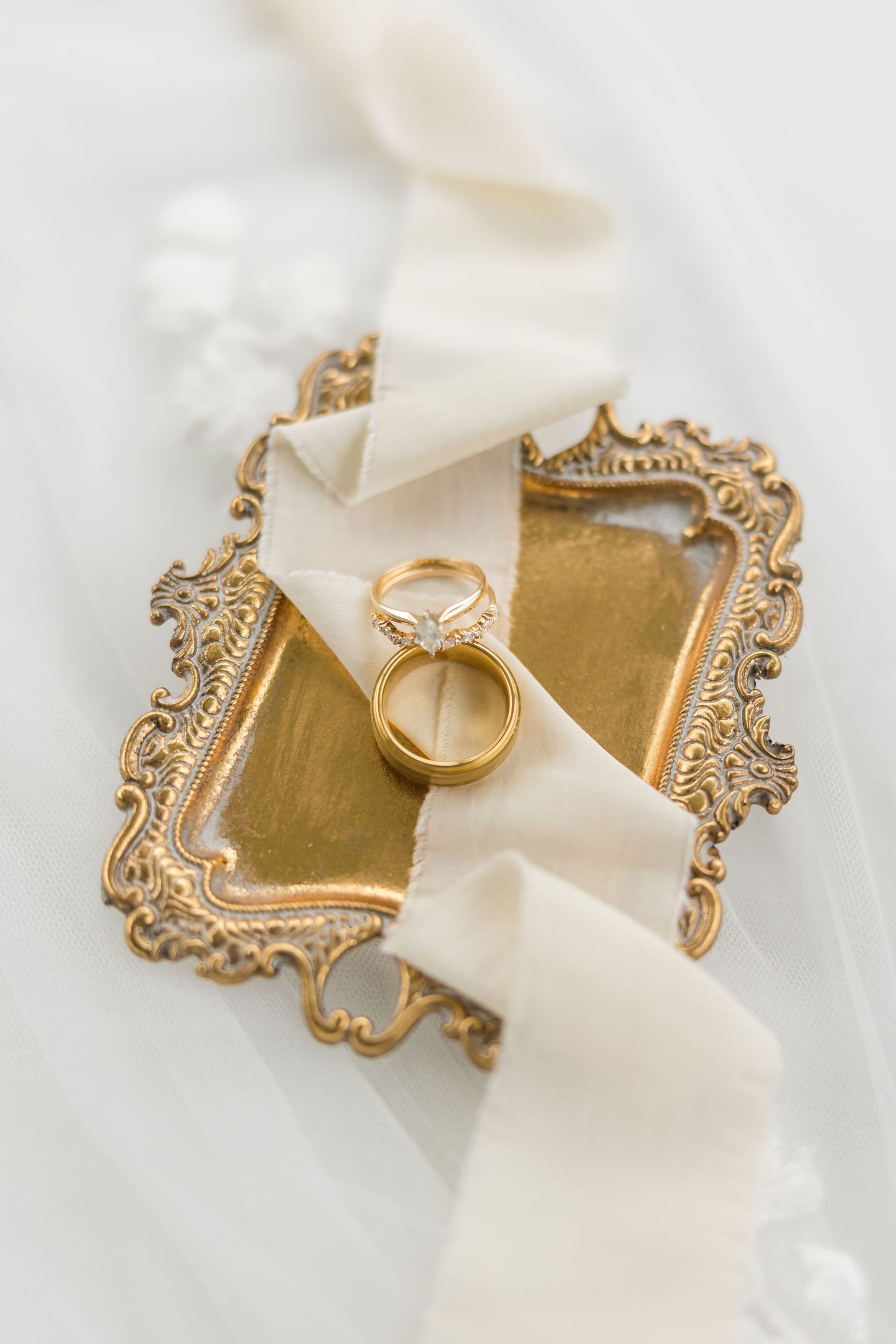 Gold wedding bands and diamond engagement ring on ornate vintage brass tray with white ribbon.