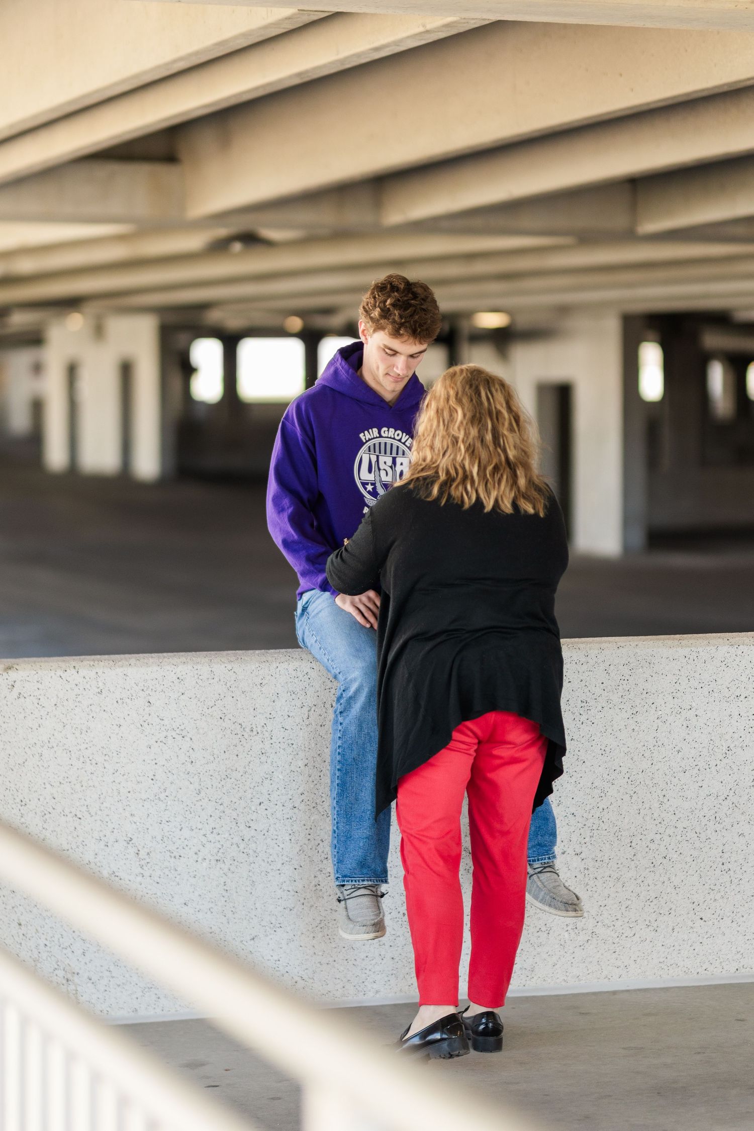 Two people talking in parking garage, one wearing purple hoodie and other in black coat with red pants.