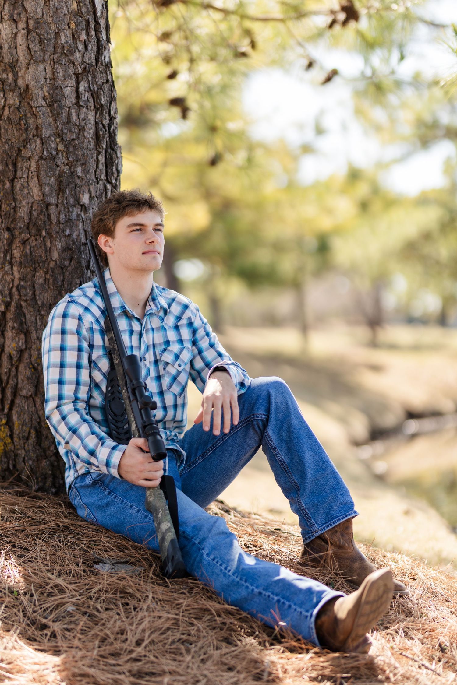 Someone wearing a plaid shirt and jeans sits casually against a tree trunk in a rustic outdoor setting.