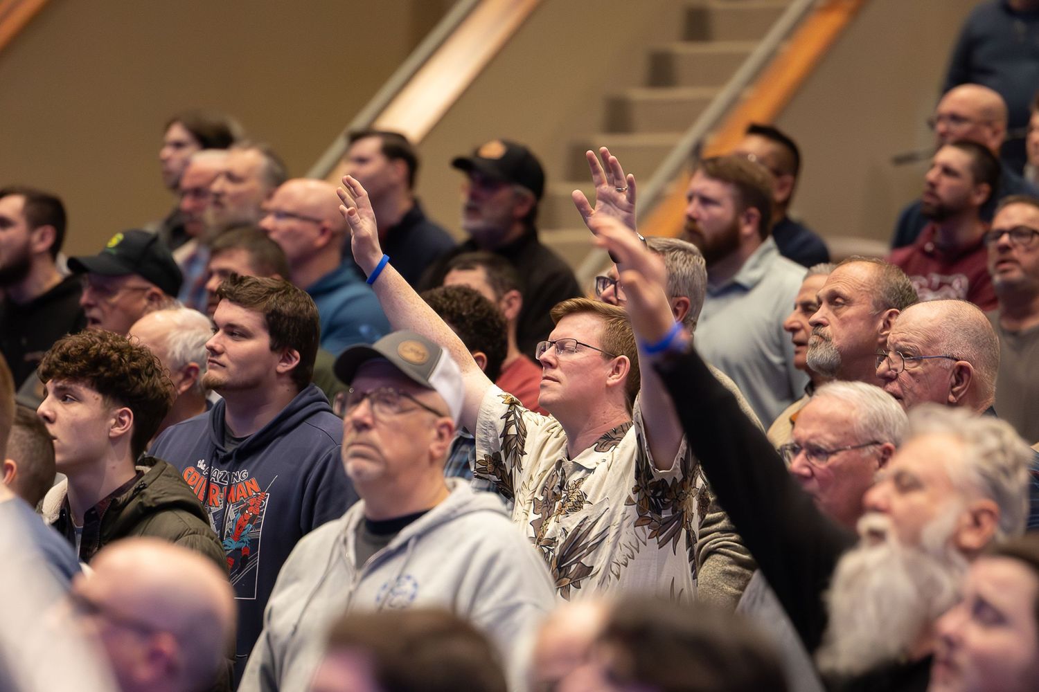 An engaged audience in a lecture hall looks upward with some members raising their hands and phones during a presentation.