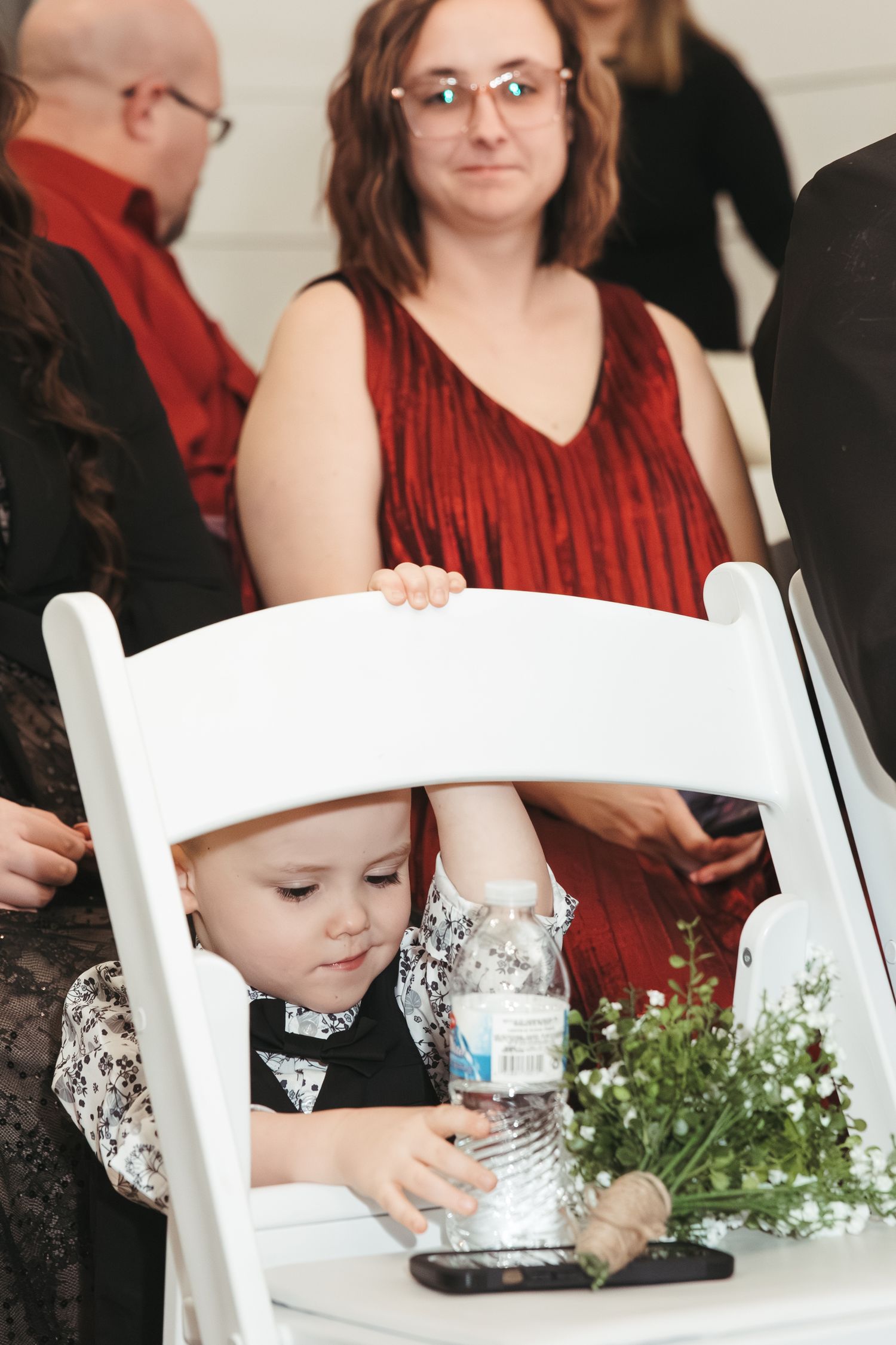 A young child in formal wear sits at a white chair during a wedding reception.